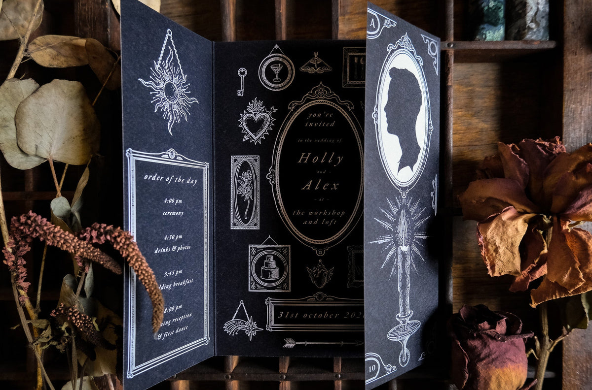 Candle and Key black gatefold invitation with white ornate detailing among dried flowers