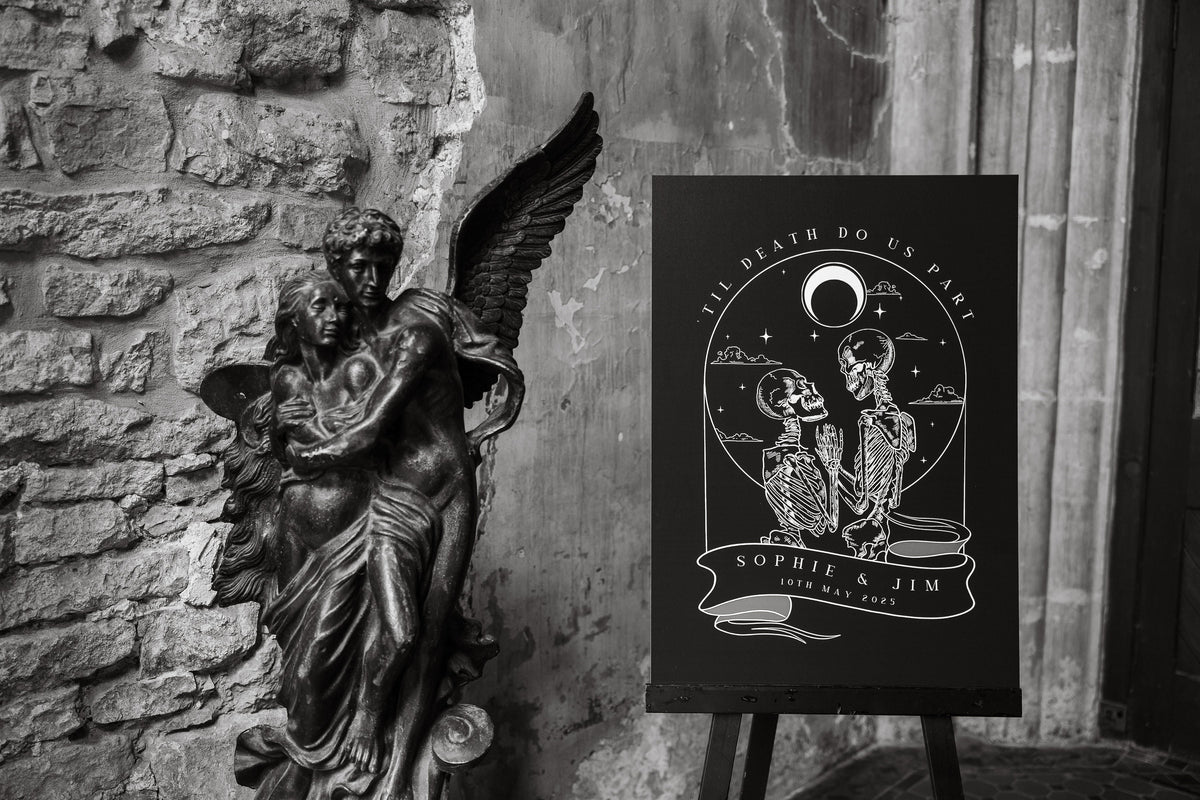 Til Death Do Us Part welcome sign with skeleton illustrations beside a stone angel statue at a real wedding