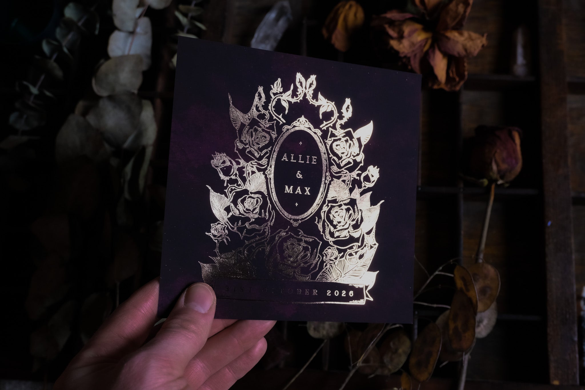 A hand holds a luxury UK save the date card with gothic dark floral design, gold foil accents, and blurred dried botanicals behind.