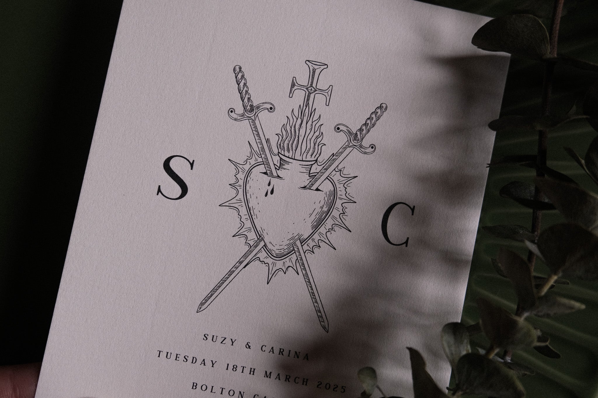 Luxury gothic wedding menu featuring a white card with dramatic tarot-style bleeding heart and crossed swords, partly veiled by leaves.