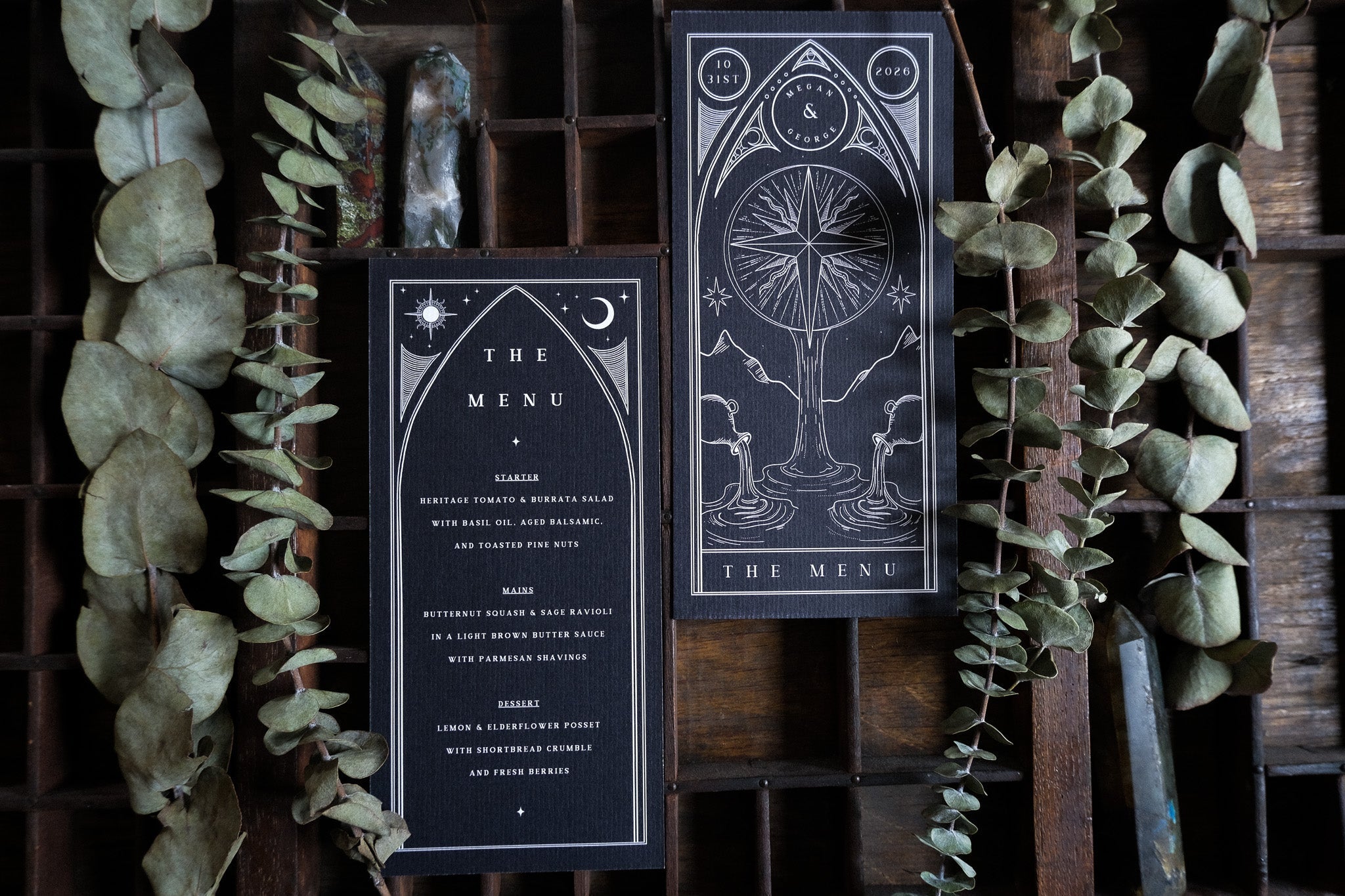 Luxury wedding menu cards in dramatic gothic black with white celestial and botanical artwork, styled with dried eucalyptus and crystals.