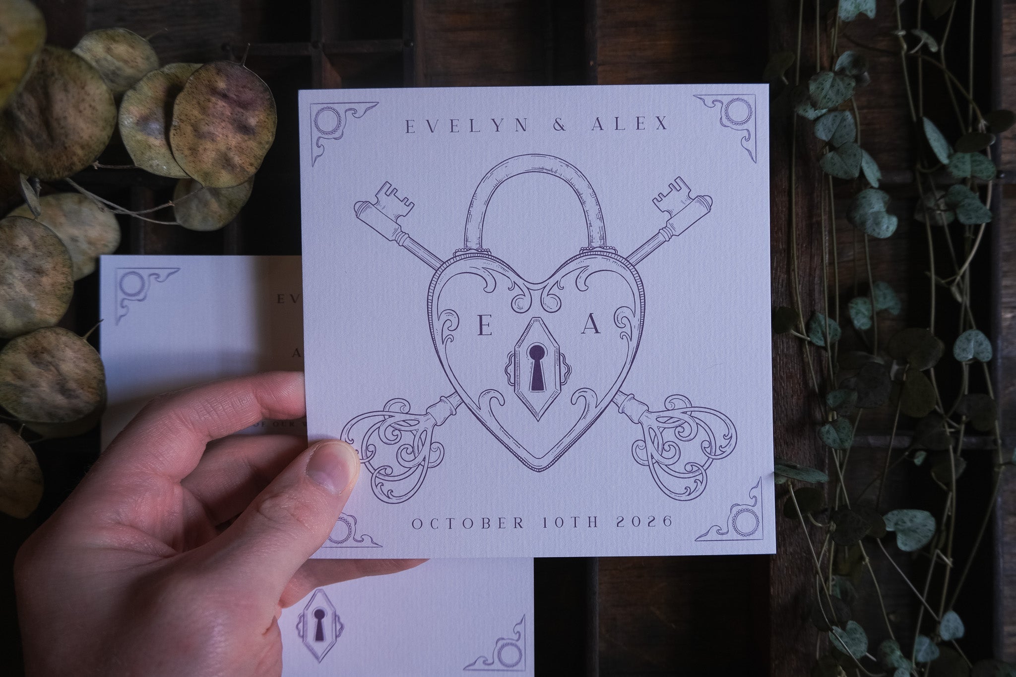 A hand presents a luxury vintage save the date card with heart padlock, ornate keys, floral accents and romantic gothic details.