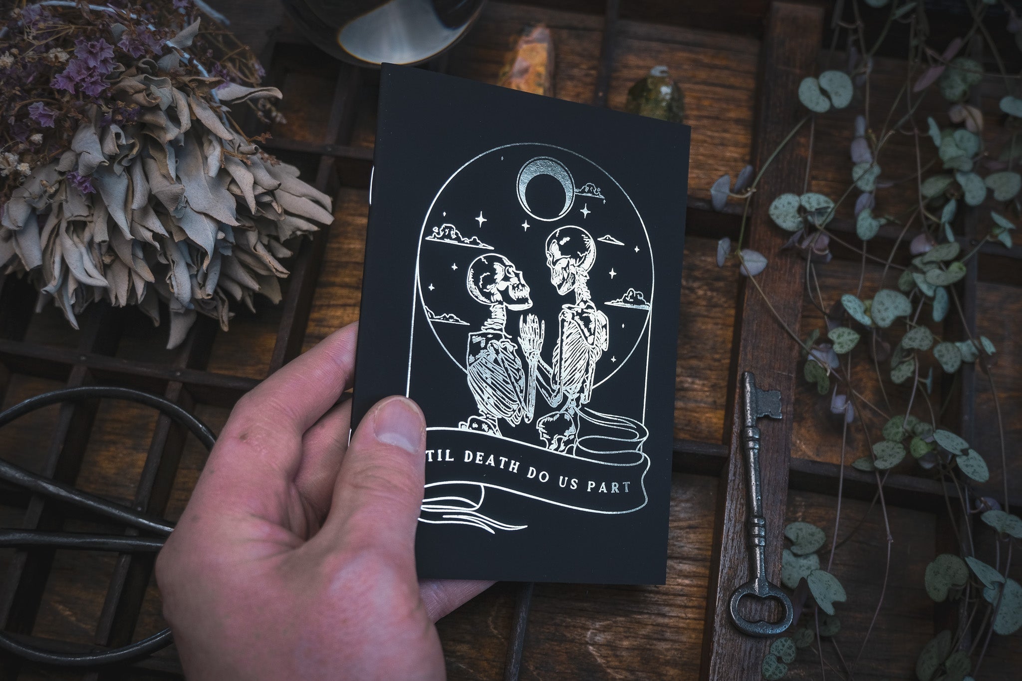 A hand holds a luxury gothic wedding vow book with dramatic gold foil skeleton and moon design, surrounded by dried flowers and vintage key.