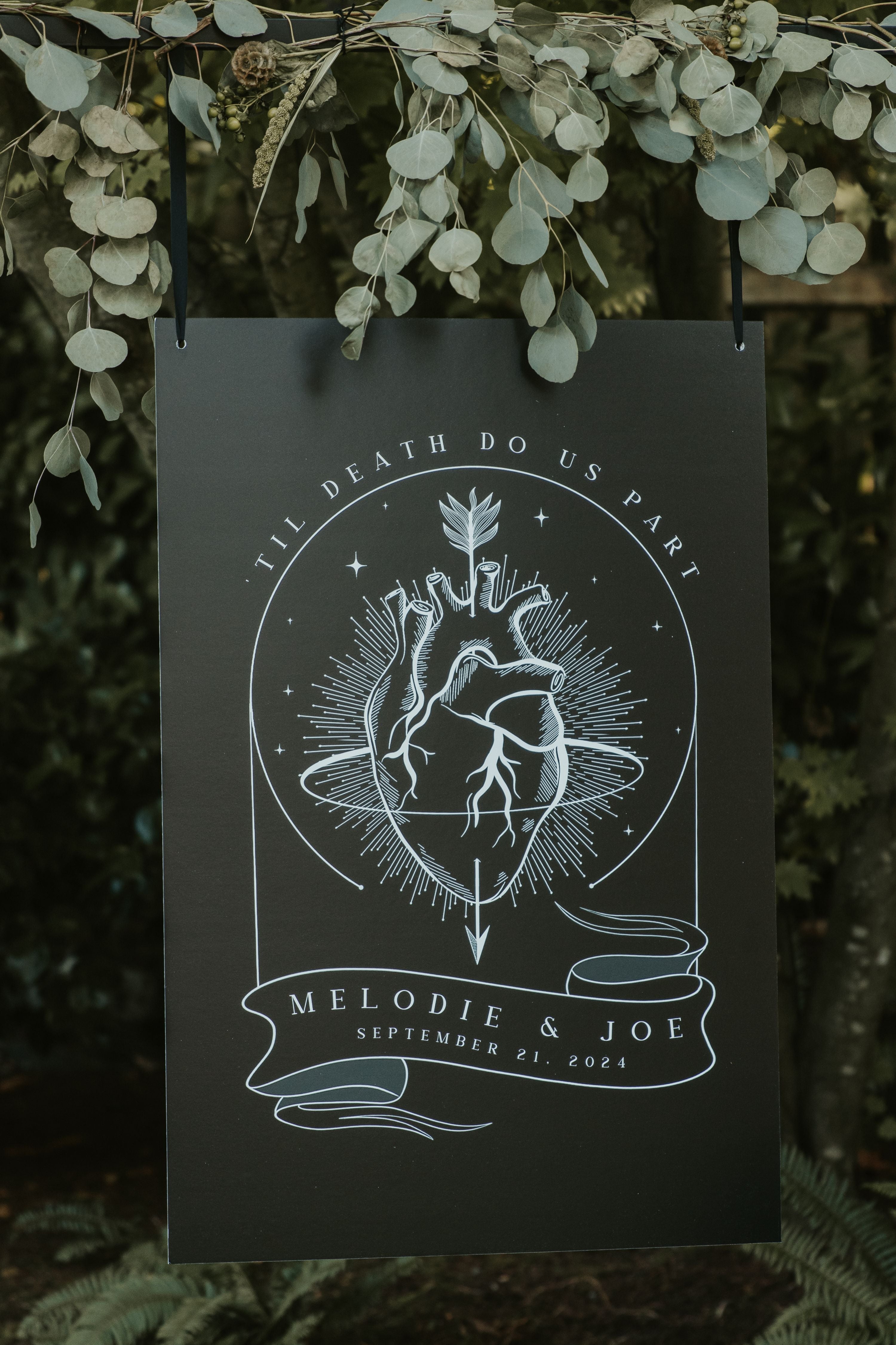 Luxury gothic wedding welcome board with mystical tarot-inspired white line art of anatomical hearts and foliage on black, set amid greenery.