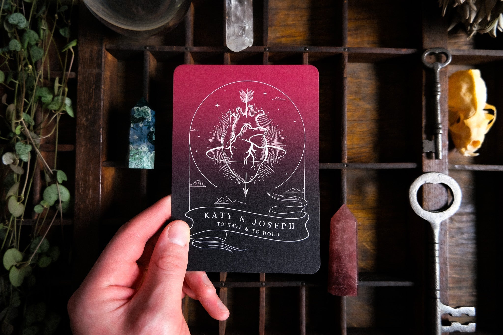A hand presents a luxury gothic A6 save the date card with an anatomical heart and celestial rays, surrounded by vintage treasures.