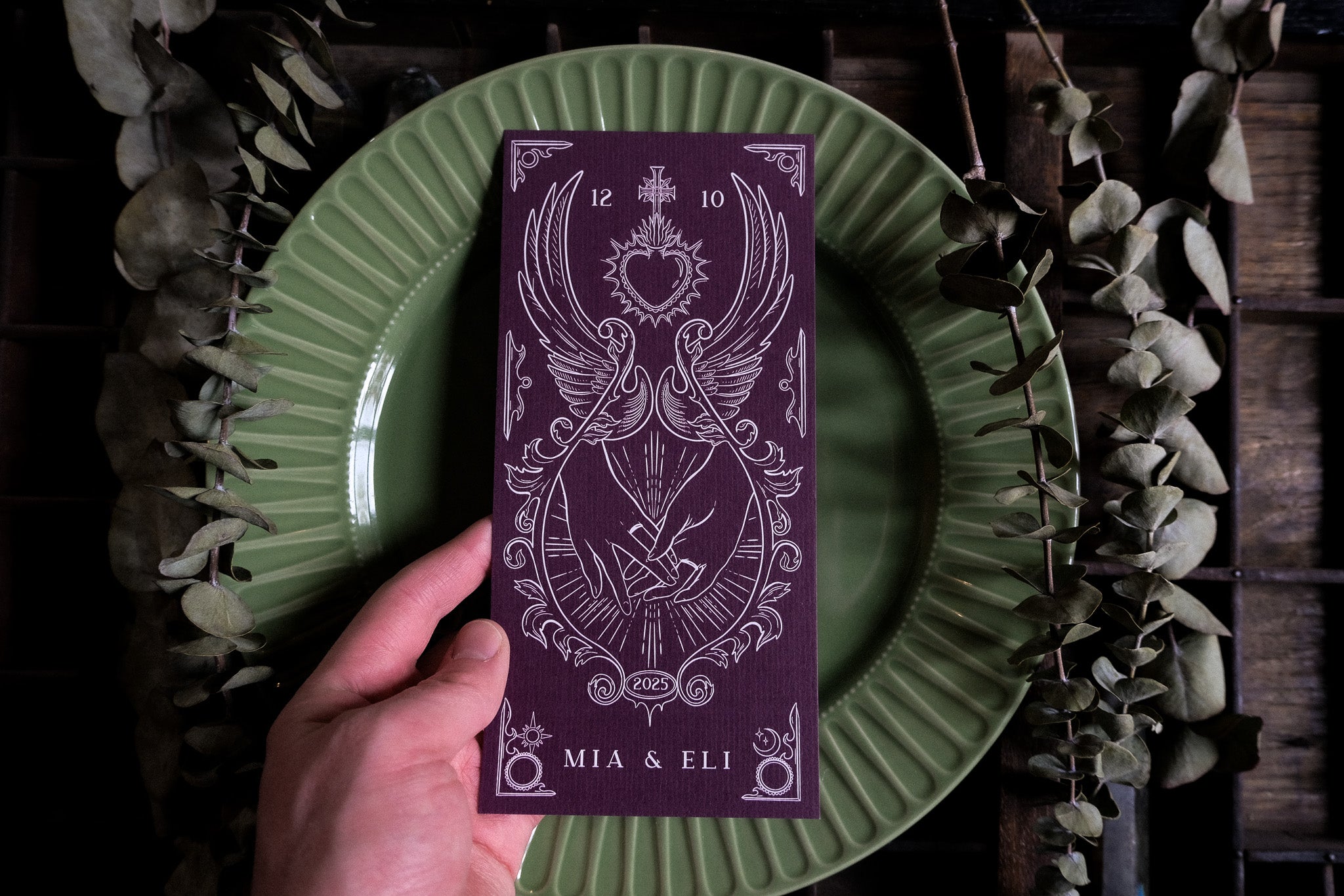 A hand displays luxury tarot-inspired wedding stationery with intricate white detailing on deep green, styled with dried eucalyptus for a gothic UK table.