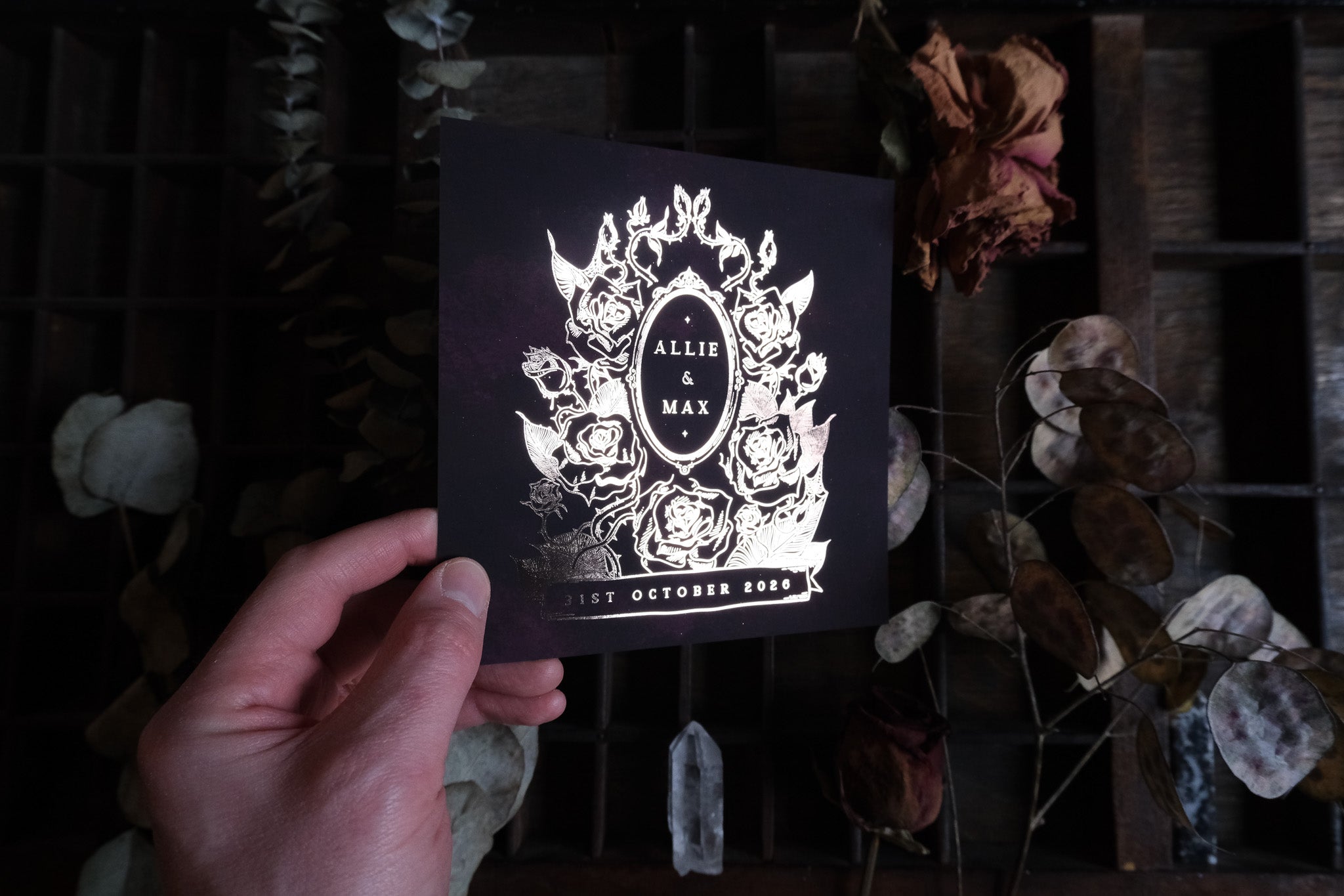 A hand displays luxury gothic wedding stationery, with gold foil floral details on dark card; dried blooms and wood create a dramatic mood.