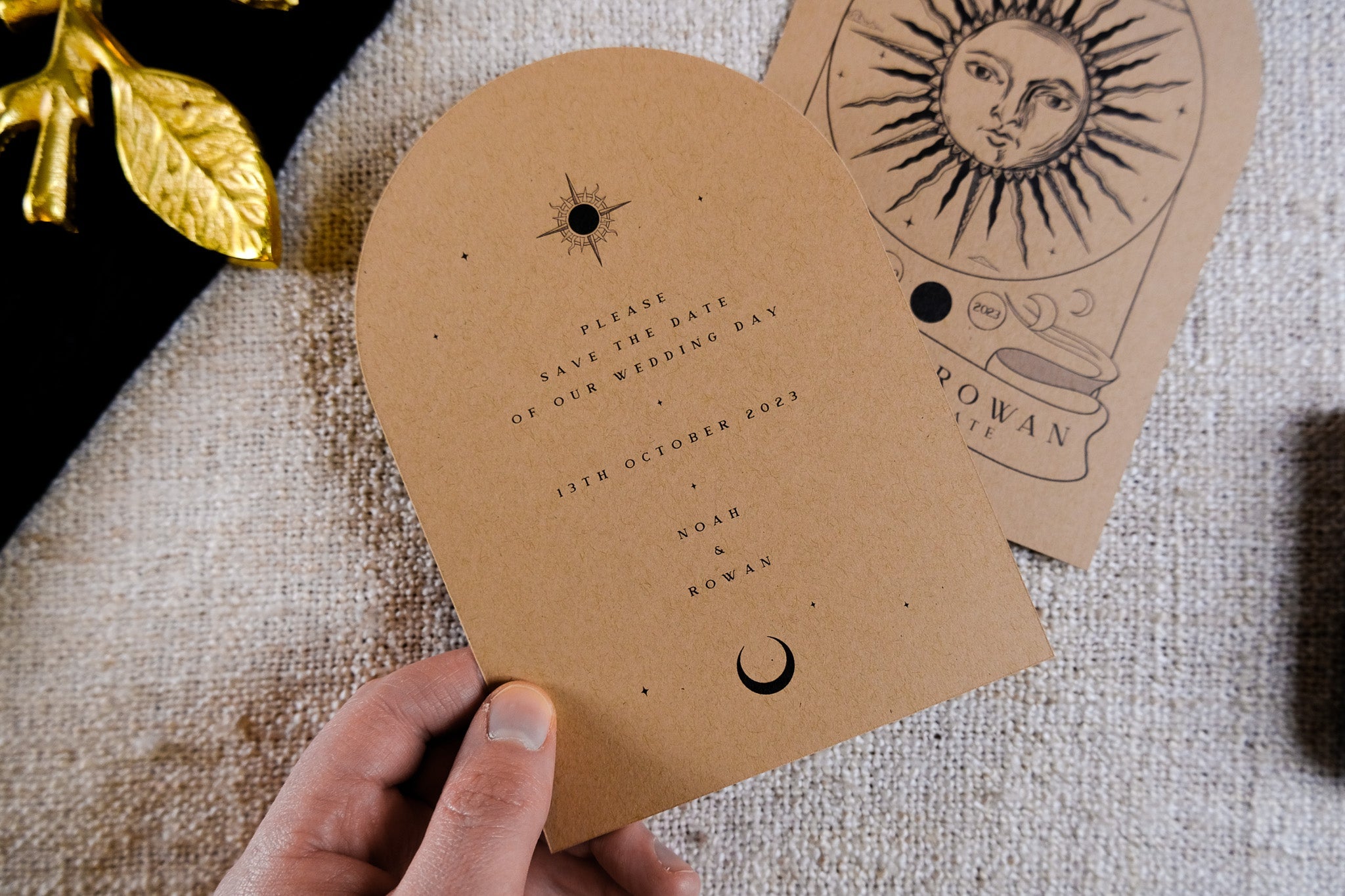 A hand displays a luxury boho wedding stationery card in kraft brown with arched shape, vintage tarot-inspired celestial motifs.