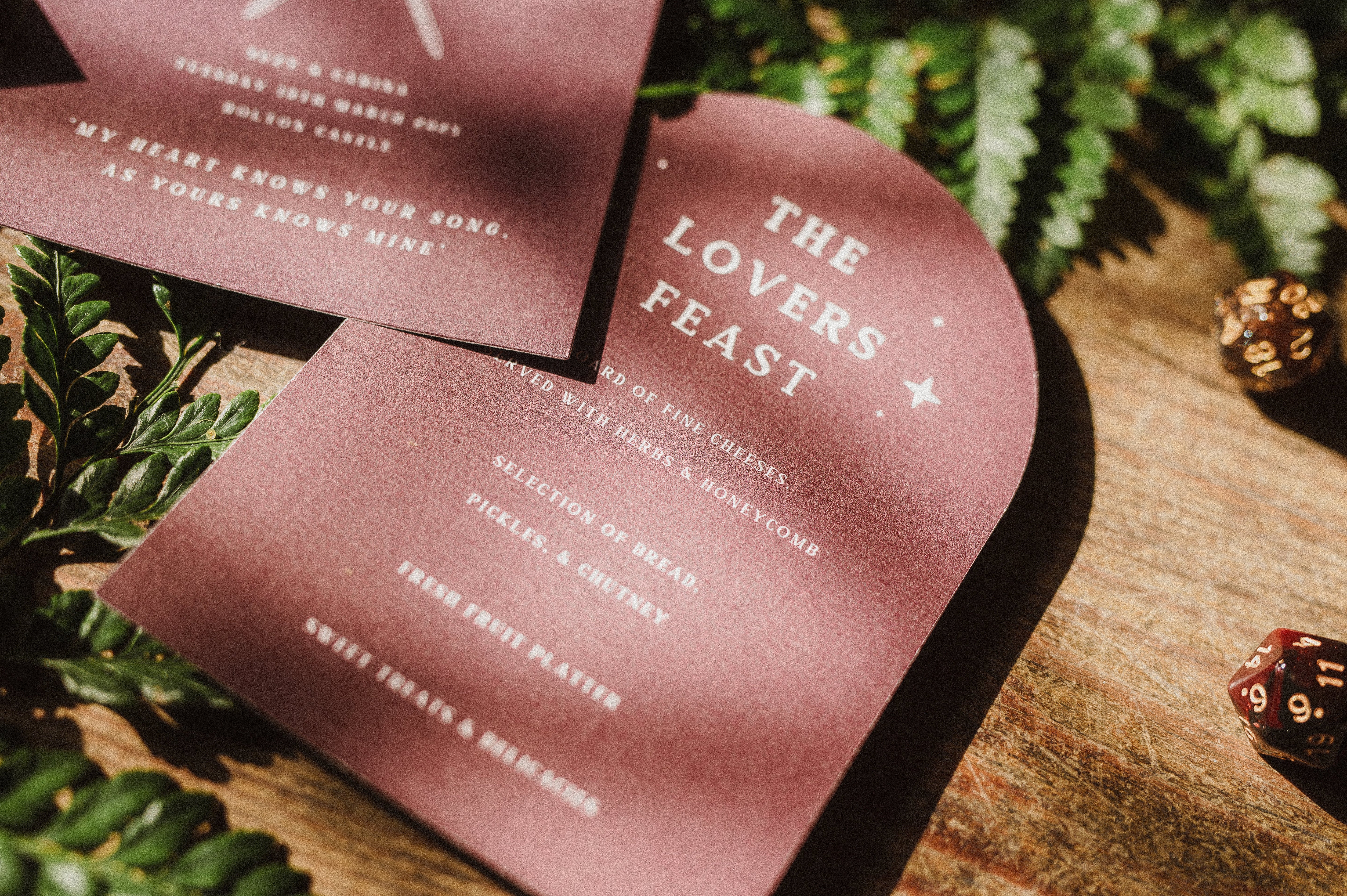 Luxury arched burgundy wedding menus with gothic bleeding heart and swords motif, set on wood with ferns and a mystical 20-sided die.