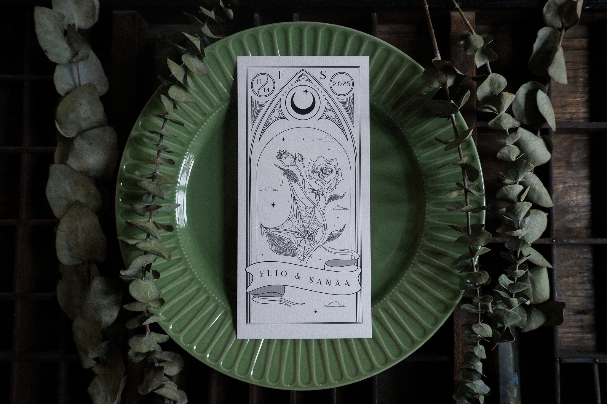 Luxury black-and-white gothic wedding menu with tarot-inspired illustrations, styled on a green plate with dried eucalyptus.