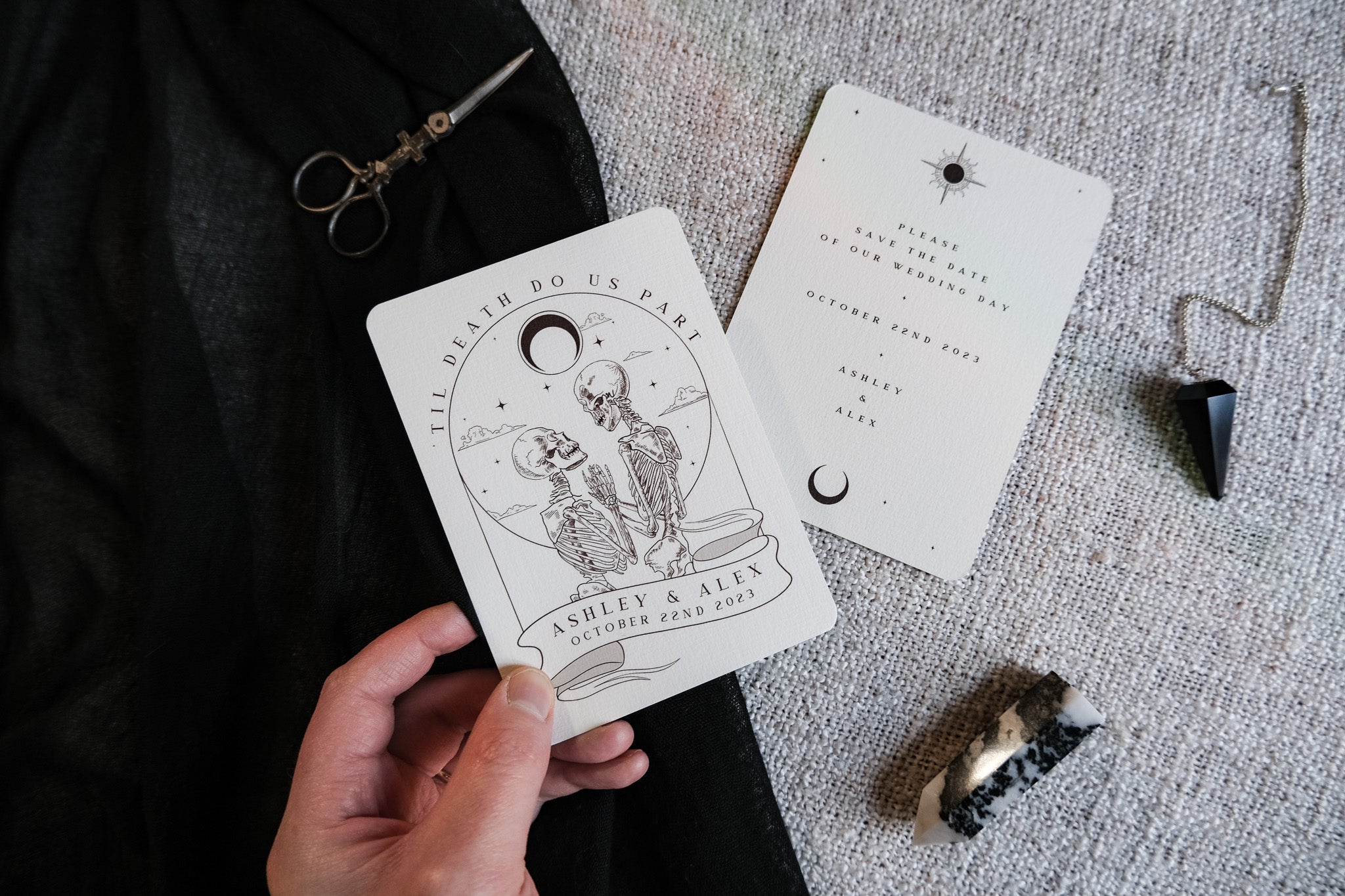 A hand holds a luxury gothic wedding save the date card with two skeletons and a moon, featuring mystical tarot-inspired artwork.