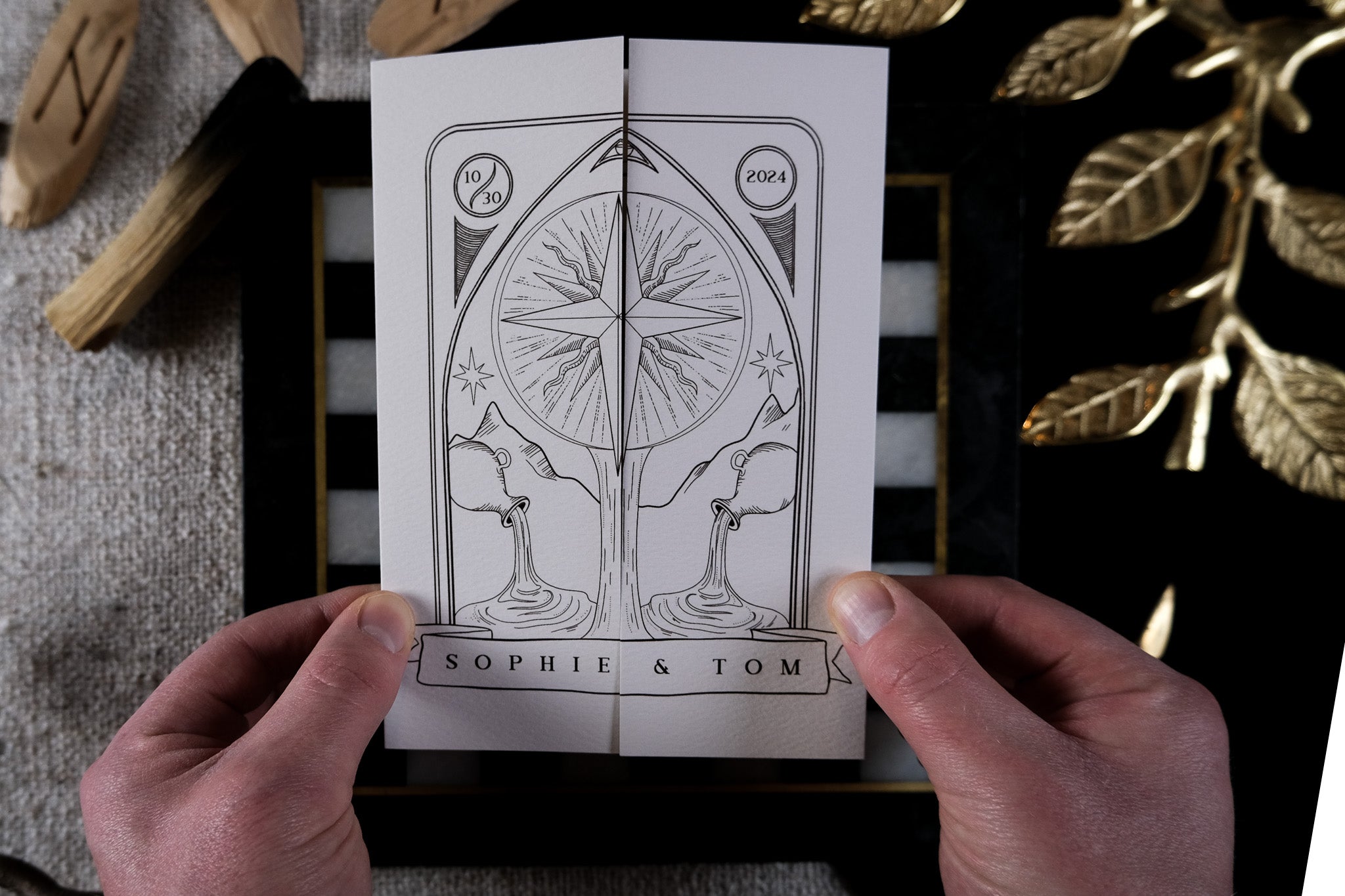 Two hands hold a luxury celestial gatefold wedding invitation with a gothic swan and star design, styled with gold leaves and wood accents.