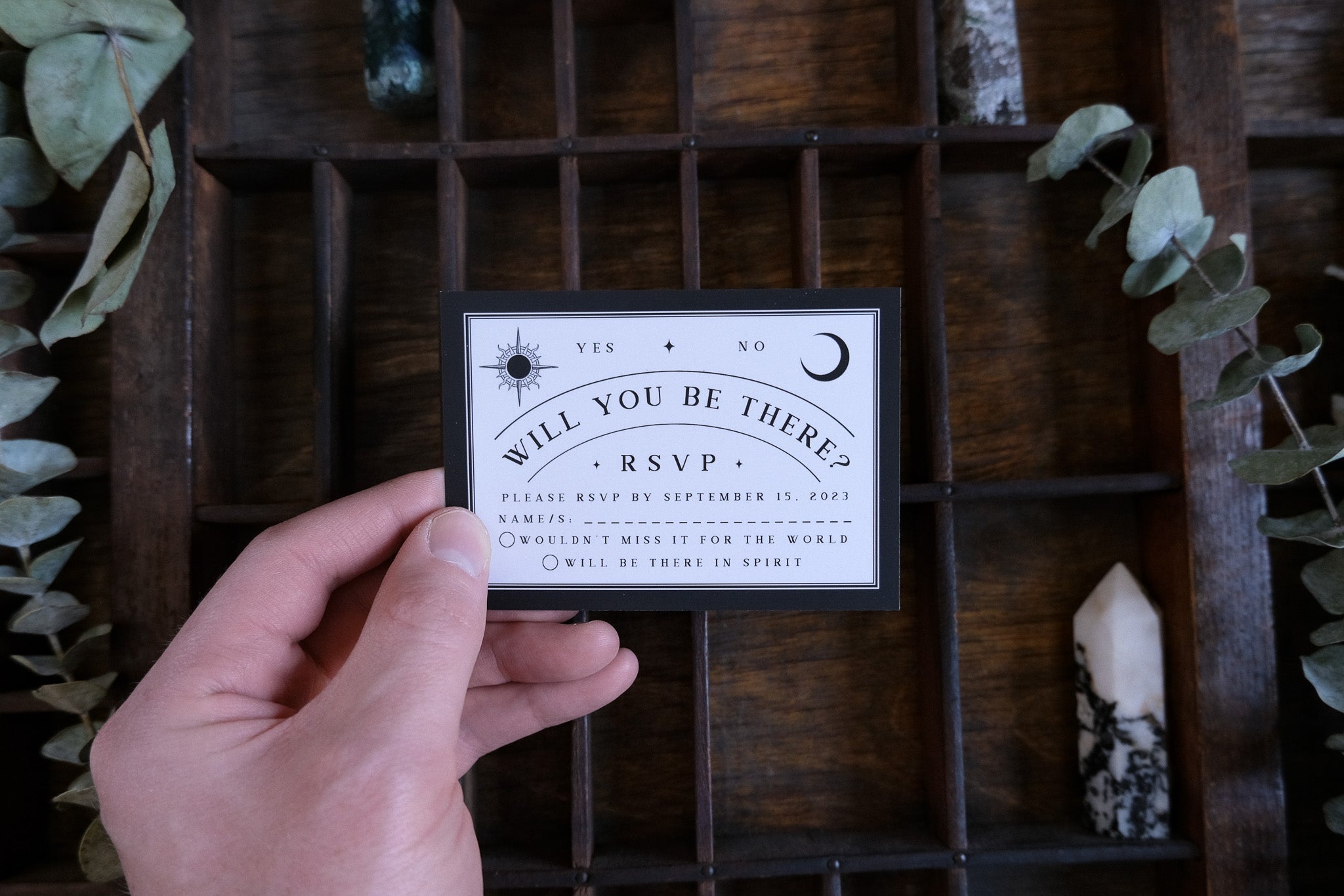 A hand displays a gothic black and white RSVP card, featuring mystical motifs, set among greenery and crystals on rustic wood.