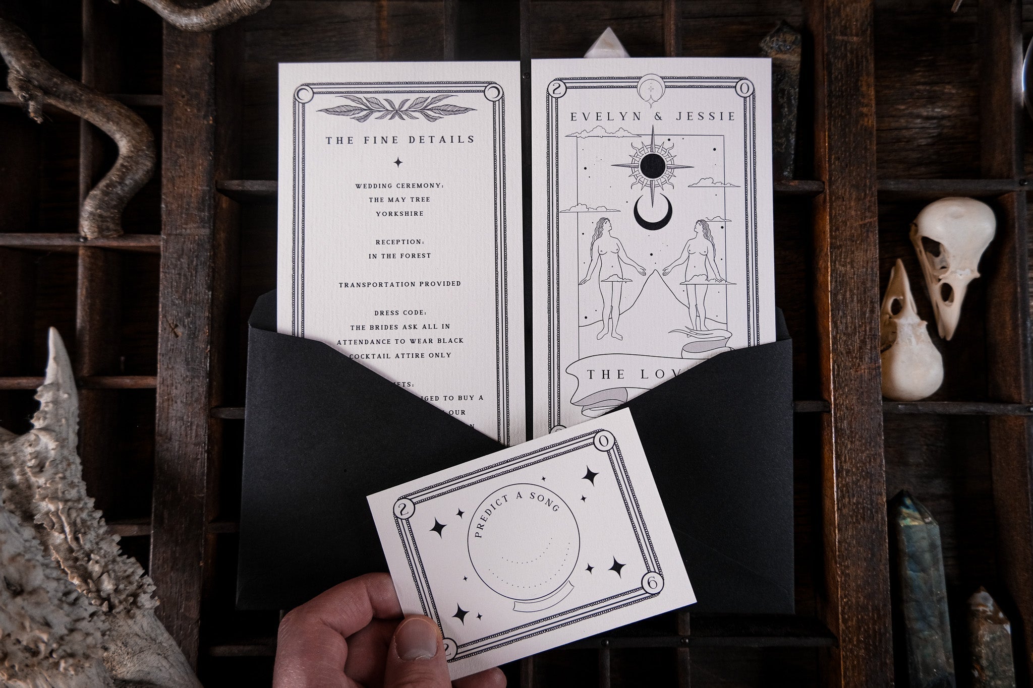 A hand holds a luxury gothic wedding invitation suite with dark romantic design over a black envelope, beside bones on a wooden tray.