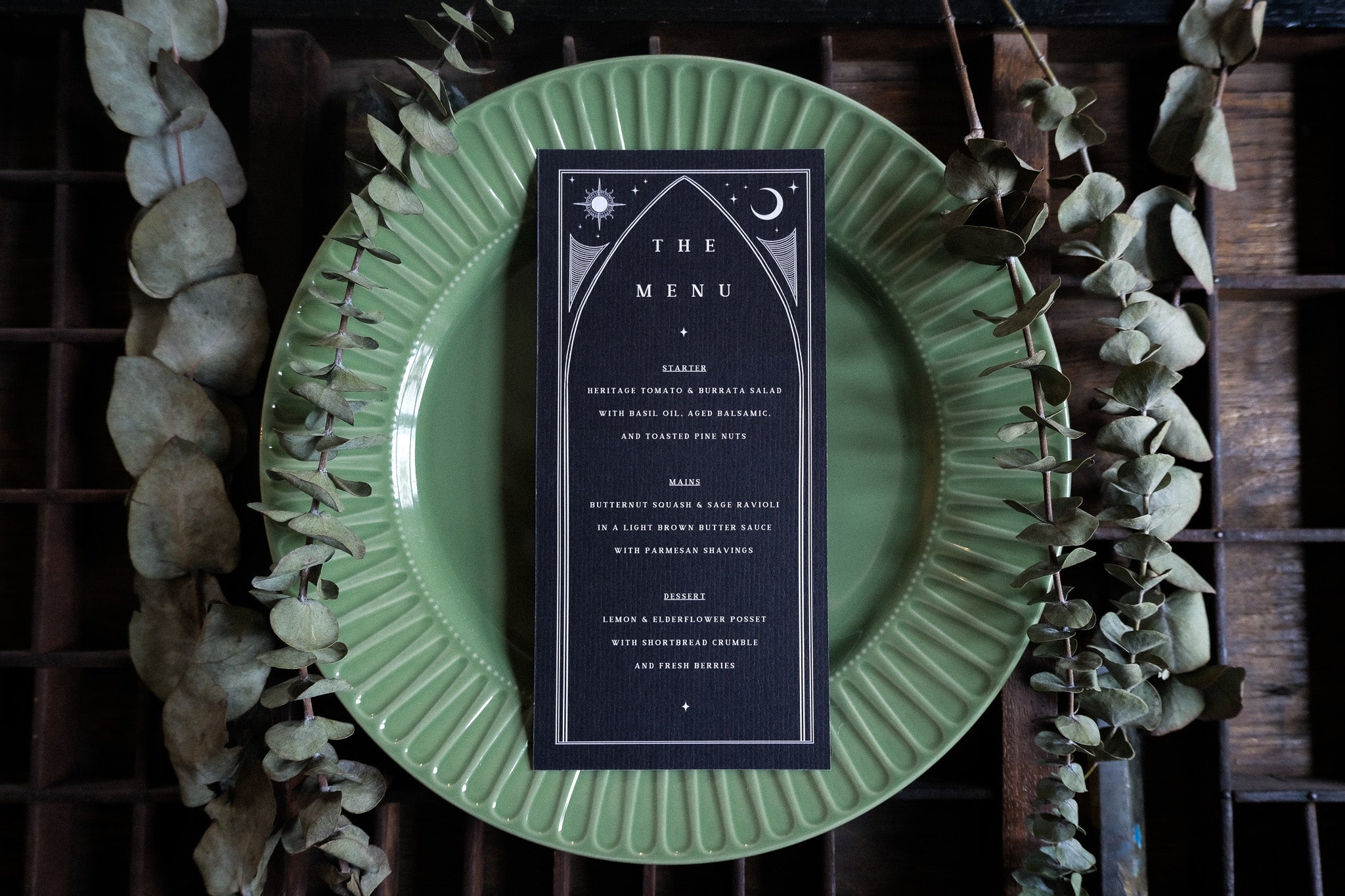 Celestial black and white wedding menu card with mystical, alternative design on green plate, rustic wood, and dried eucalyptus.