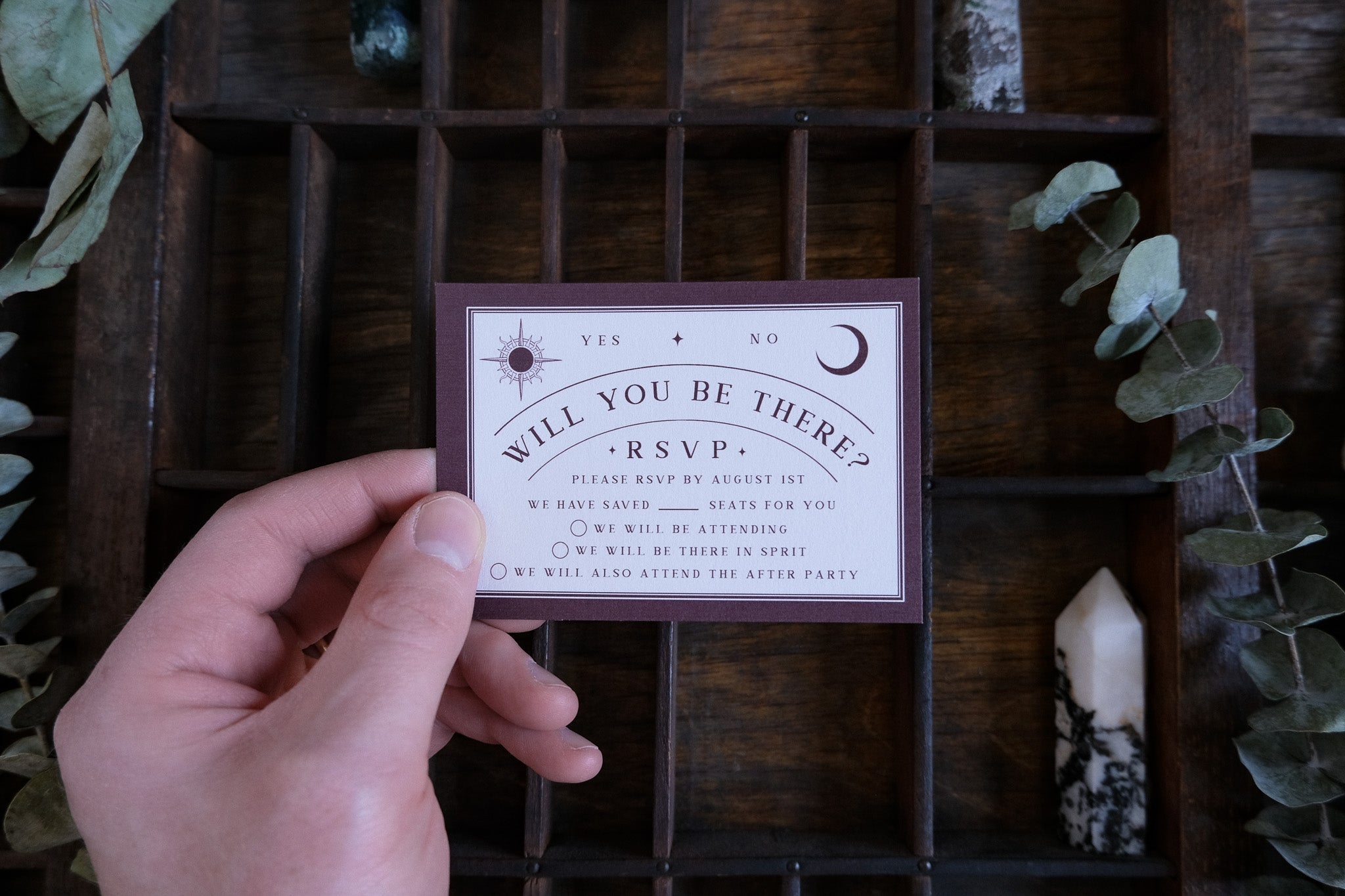 A hand presents a gothic-inspired RSVP card with vintage motifs, set among crystals, greenery and stones for mystical UK wedding stationery.