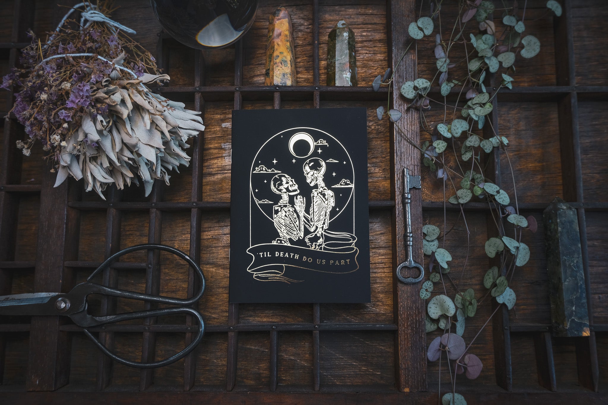 Luxury black wedding vow book with foiled skeletal lovers under a moon, styled among dried flowers and candles for a gothic UK display.