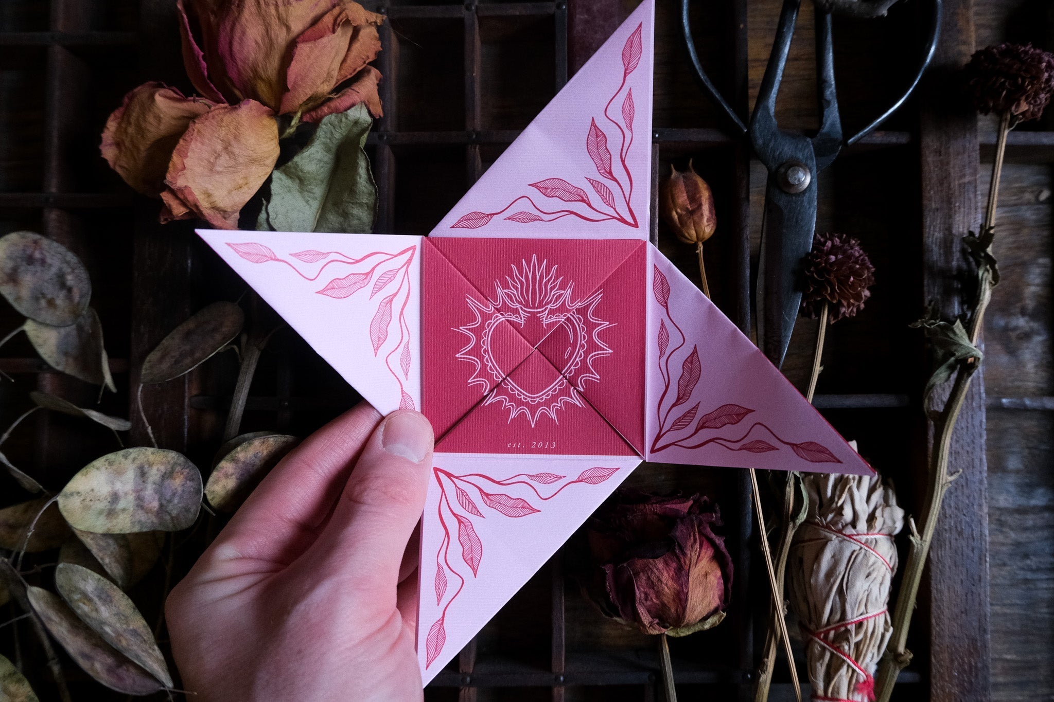 A hand holds a luxury celestial pink and red vintage folding wedding card with romantic heart motifs, set among dried flowers and leaves.