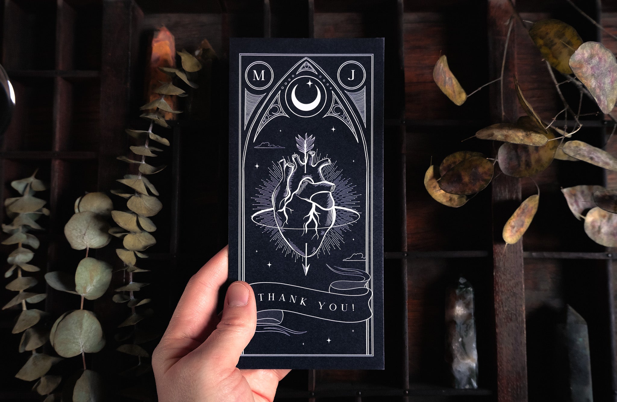 A hand holds a luxury personalised UK wedding thank you card with an anatomical heart and celestial motifs, in a vintage boho setting.