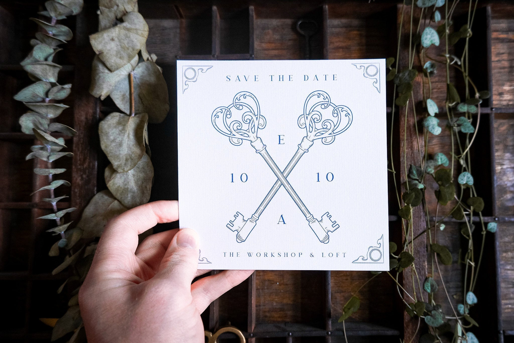 A hand holds a romantic, vintage-inspired luxury save the date card with ornate skeleton keys, set on rustic wood and dried foliage.