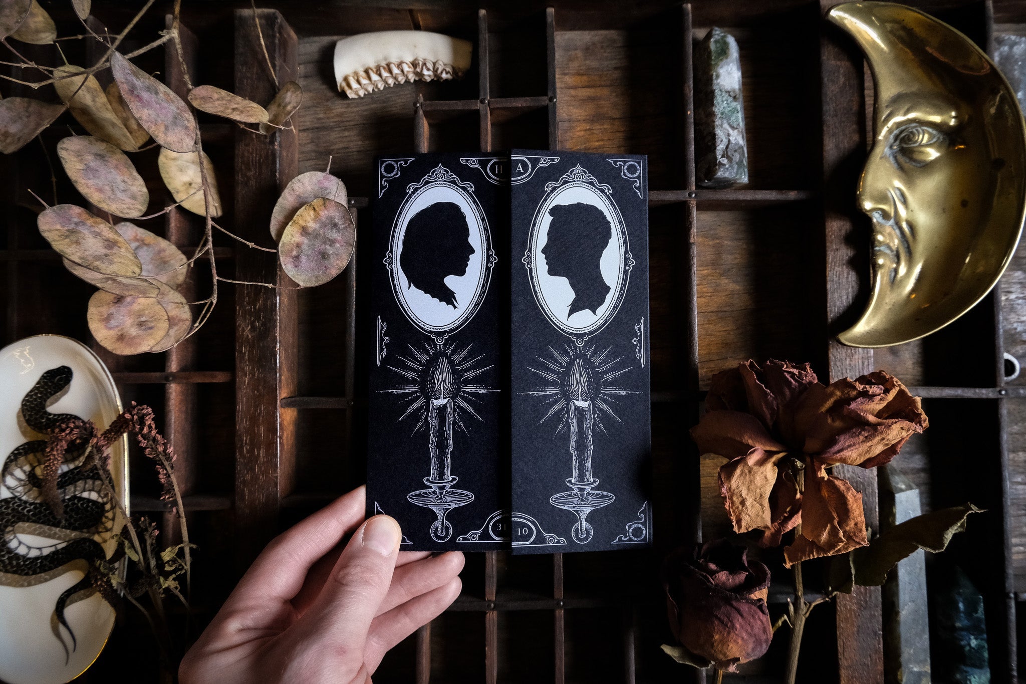 A hand holds two luxury black gatefold gothic wedding invitations with white silhouette portraits and candle art, styled among dried leaves, roses, crystals, a celestial brass moon mask and vintage accents.