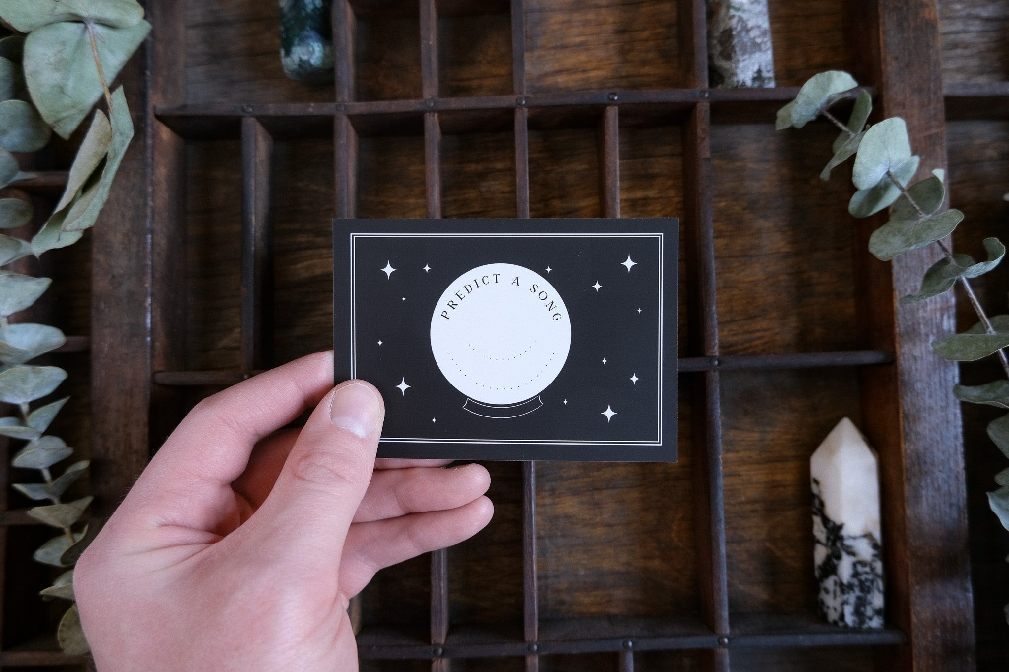 A hand holds a gothic RSVP card with a white celestial design. Crystals, foliage, and wood lend mystical luxury—UK wedding stationery.