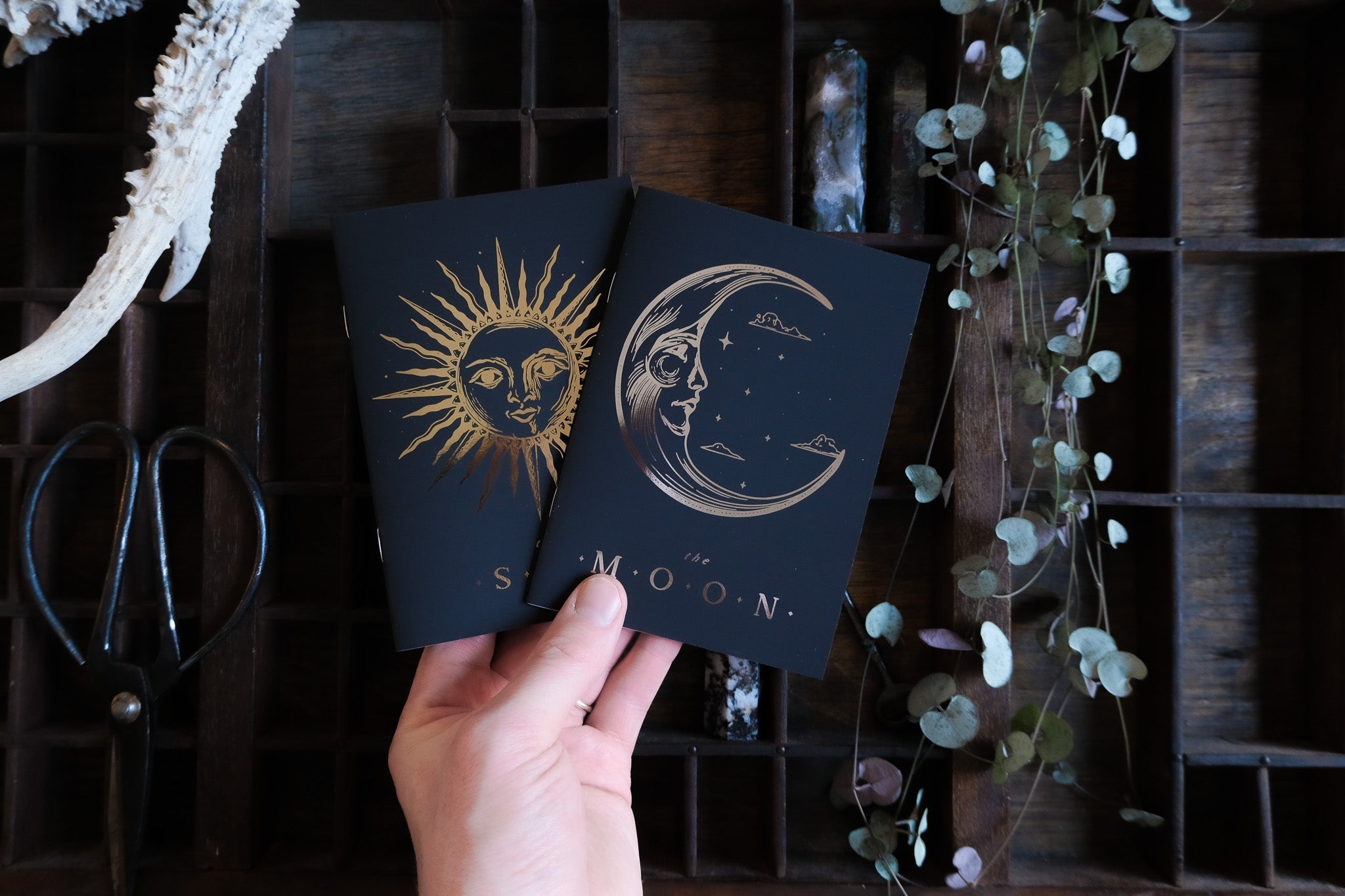 A hand displays luxury gold and silver foiled gothic wedding vow books with celestial motifs; boho props and vintage accents surround.