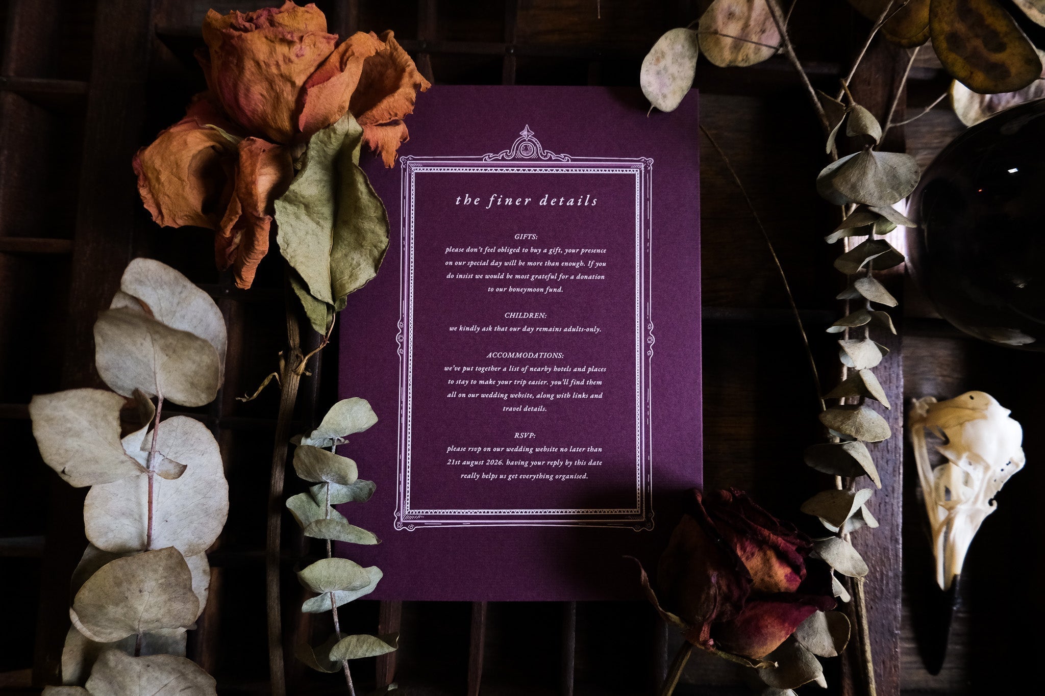 Luxury gothic gatefold wedding stationery in deep tones, styled with dried roses, eucalyptus, and a bird skull on dark wood.