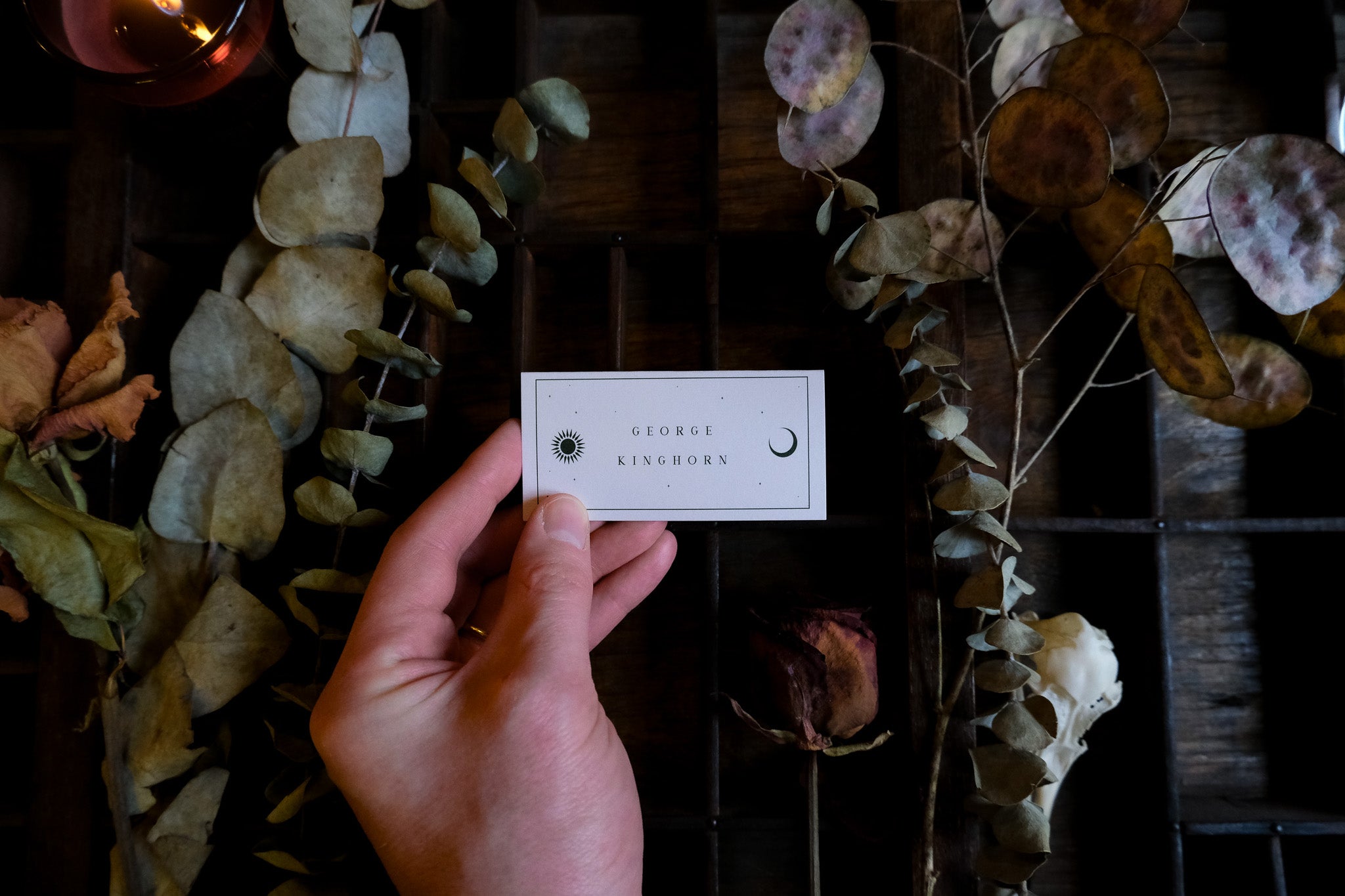 A luxury celestial wedding placecard with minimalistic sun and moon motifs, set on a dark wood among dried florals for a gothic vibe.