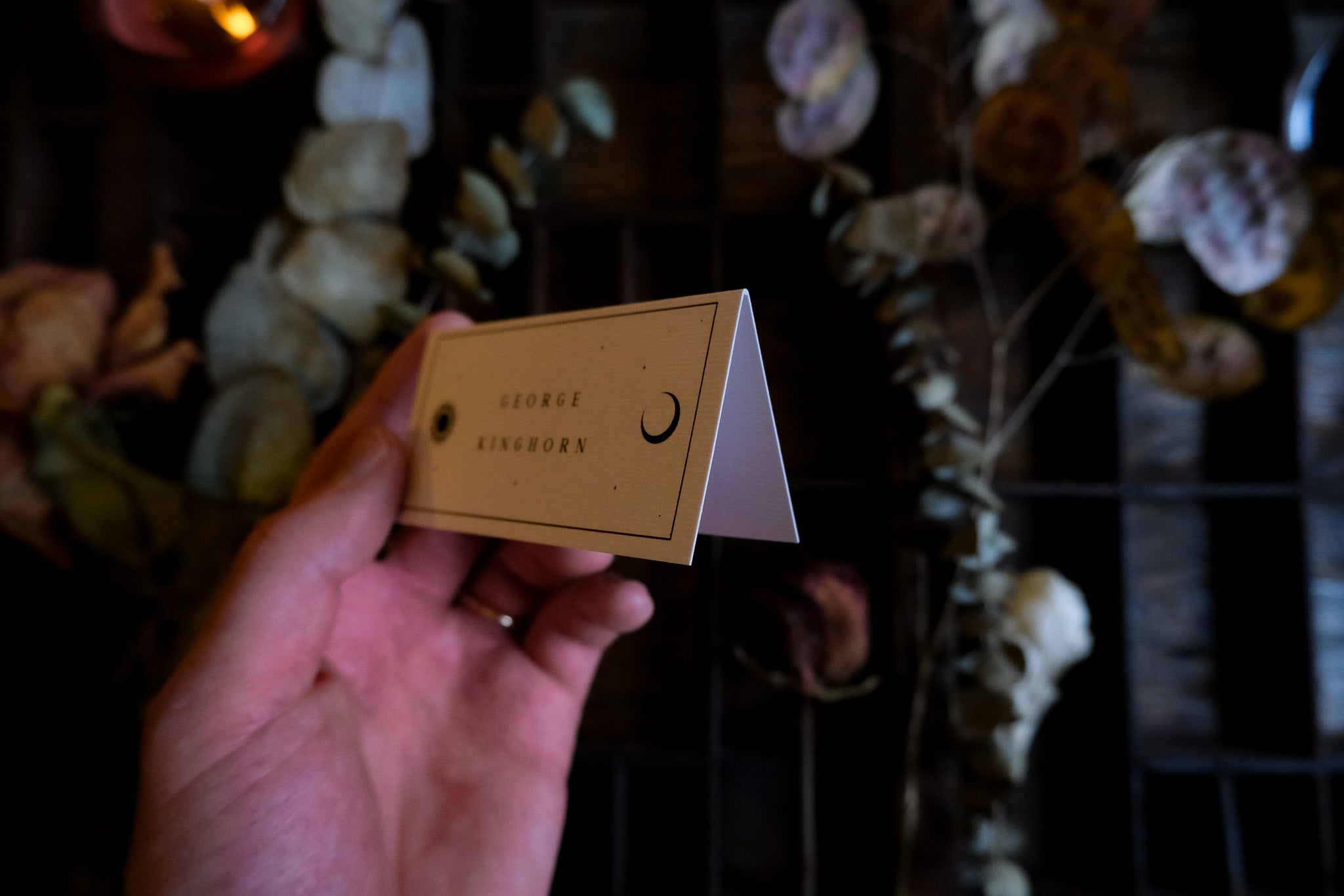 A hand holds a folded beige celestial wedding placecard with a crescent moon, in a romantic, luxury UK setting with dried florals.