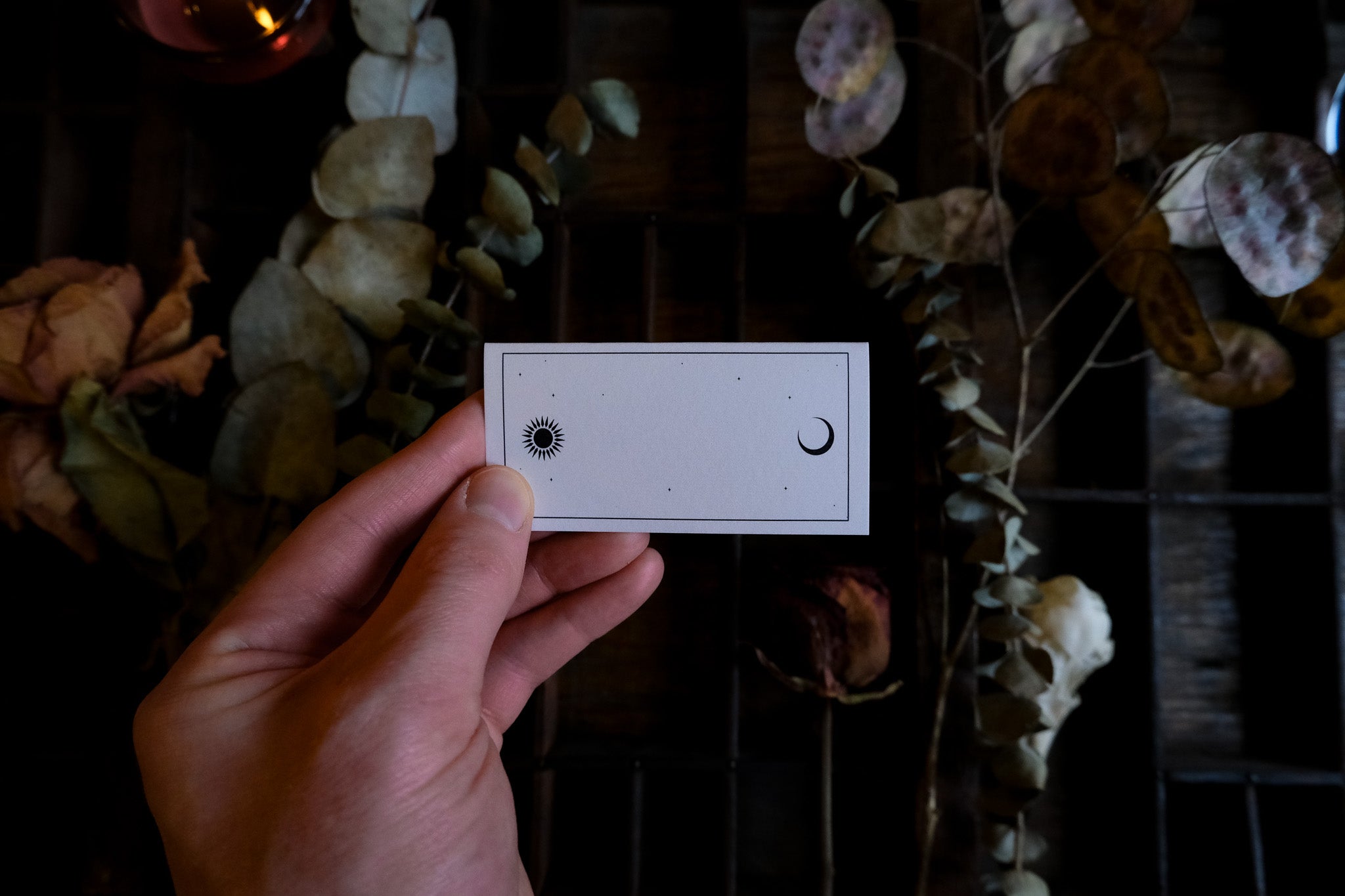 A hand holds a luxury celestial place card with black sun and crescent moon, dried foliage behind; dramatic gothic wedding stationery.