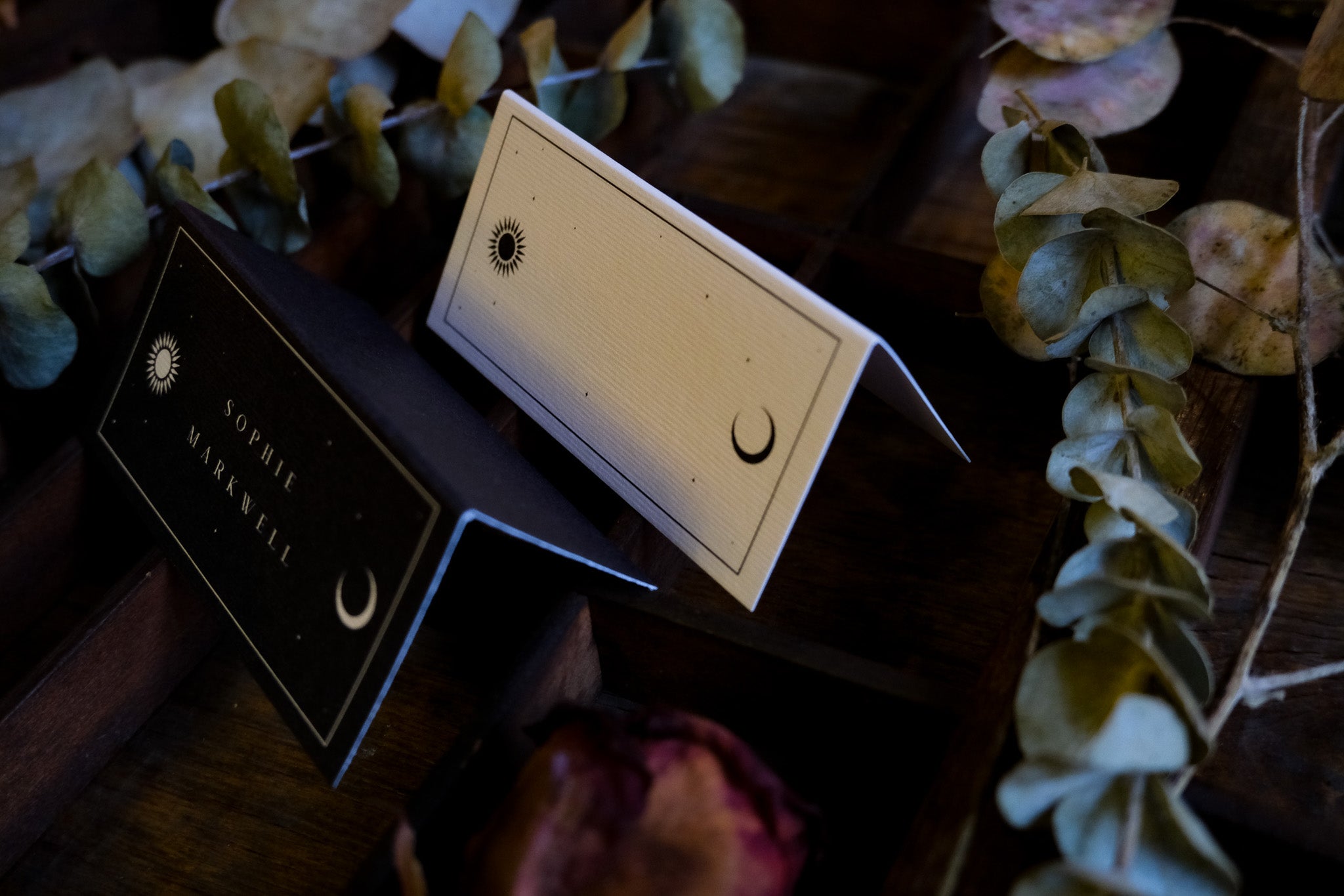 Luxury celestial wedding place cards, one black and one white, with mystical designs on wood and dried botanicals; gothic, alternative style.