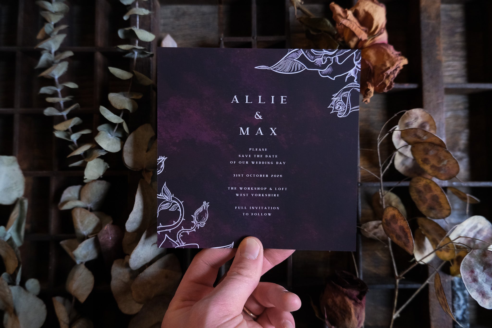 A hand presents luxury UK gothic wedding stationery, black with white florals and foil detail, styled on rustic wood with dried botanicals.