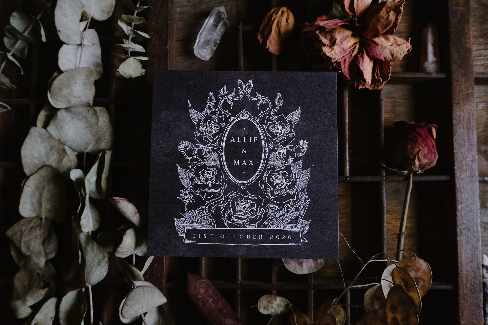 Luxury gothic save the date card with dark floral design, dried flowers, and rustic wood backdrop; dramatic UK wedding stationery.