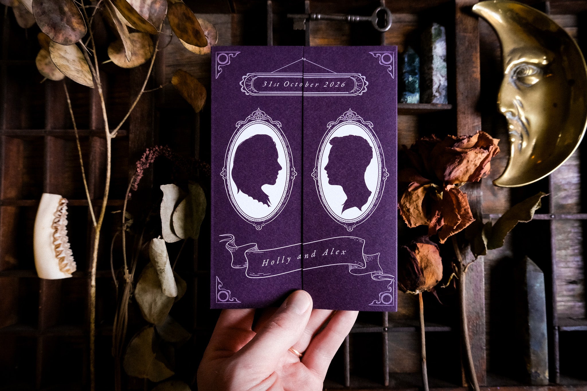 A hand holds a luxury gothic gatefold wedding invitation with bespoke silhouette art, set amid dried botanicals, bone, and celestial decor.