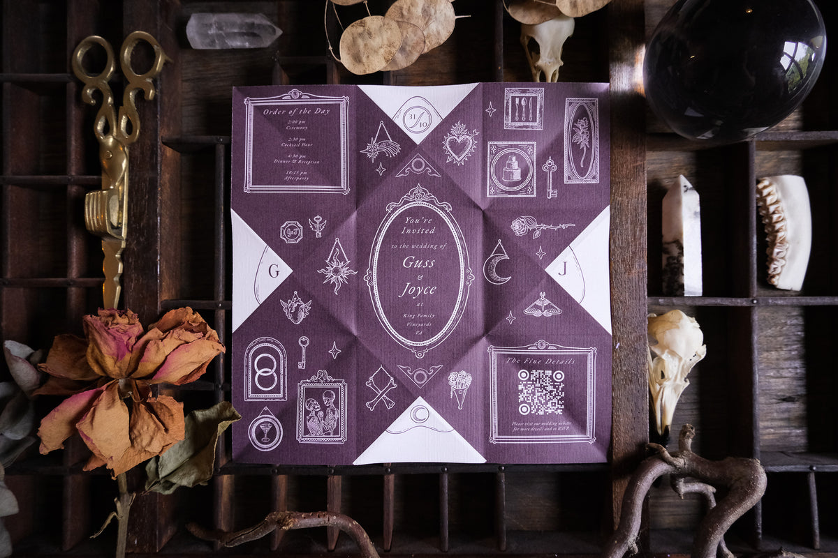A Victorian puzzle purse wedding invitation in cream and black with hand folding is styled among vintage keys, dried flowers and curios.