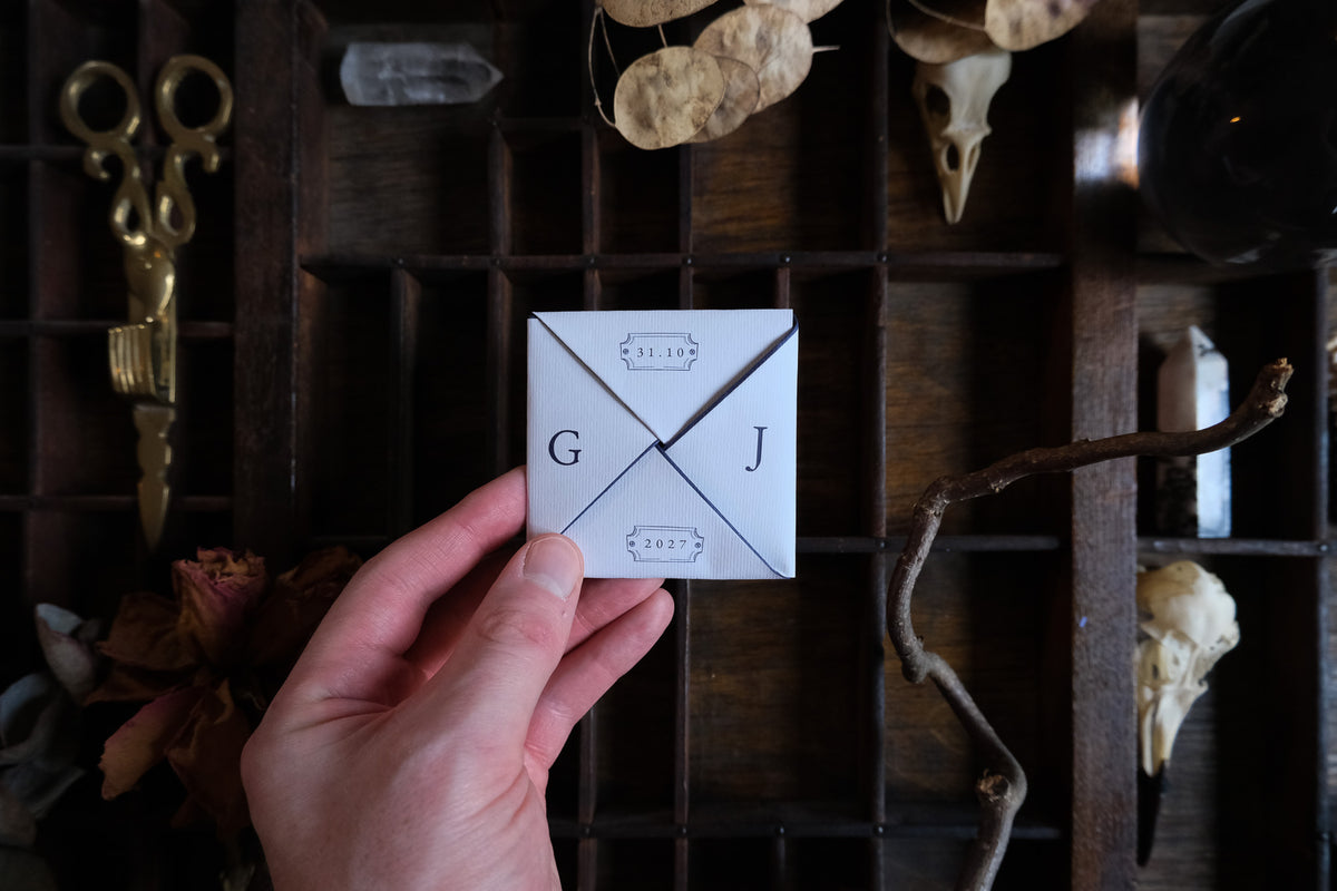 A hand displays a Victorian puzzle purse wedding invitation with candle and key illustrations on cream paper, set against a wooden organiser with scissors, dried flowers and animal skulls.