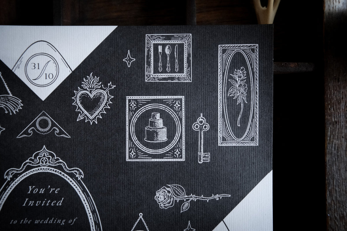 A folded black wedding invitation with white Victorian-style illustrations, photographed on a neutral tabletop.