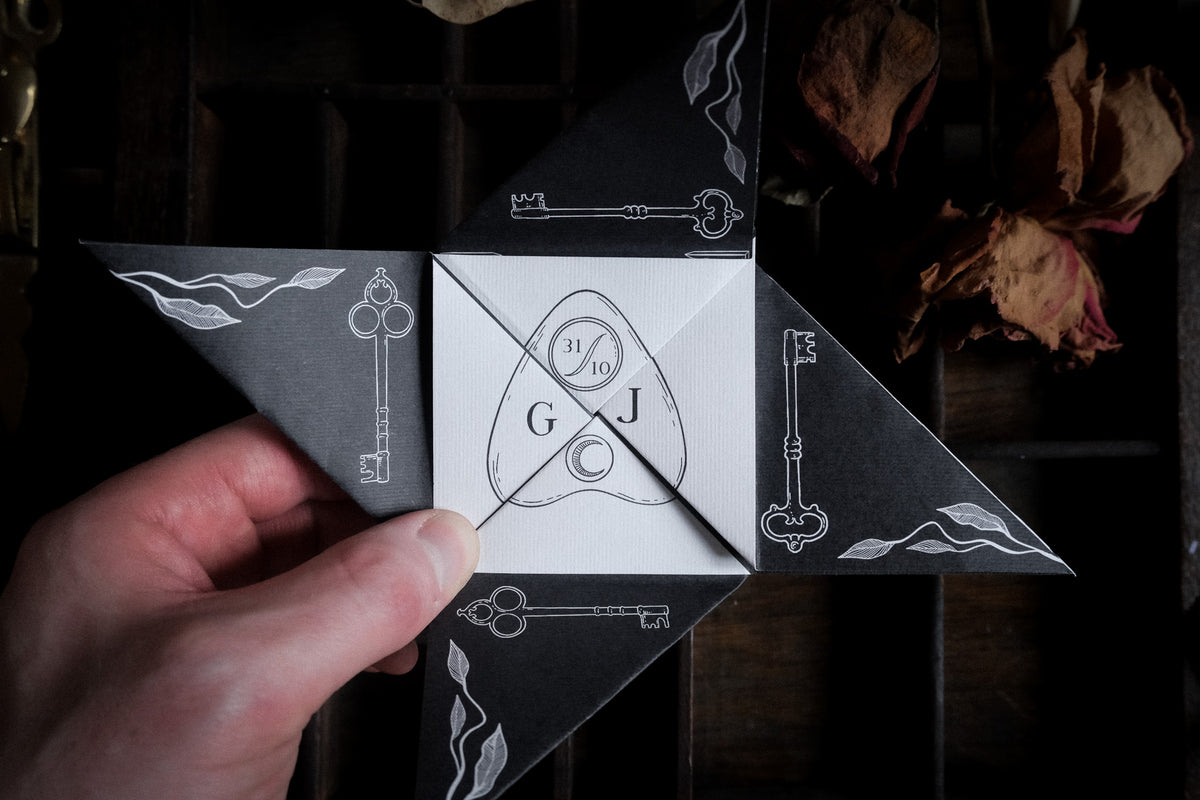 A hand holds a black and white Victorian puzzle purse wedding invitation featuring keys, flames, a crescent moon and initials, with dried roses in the background.