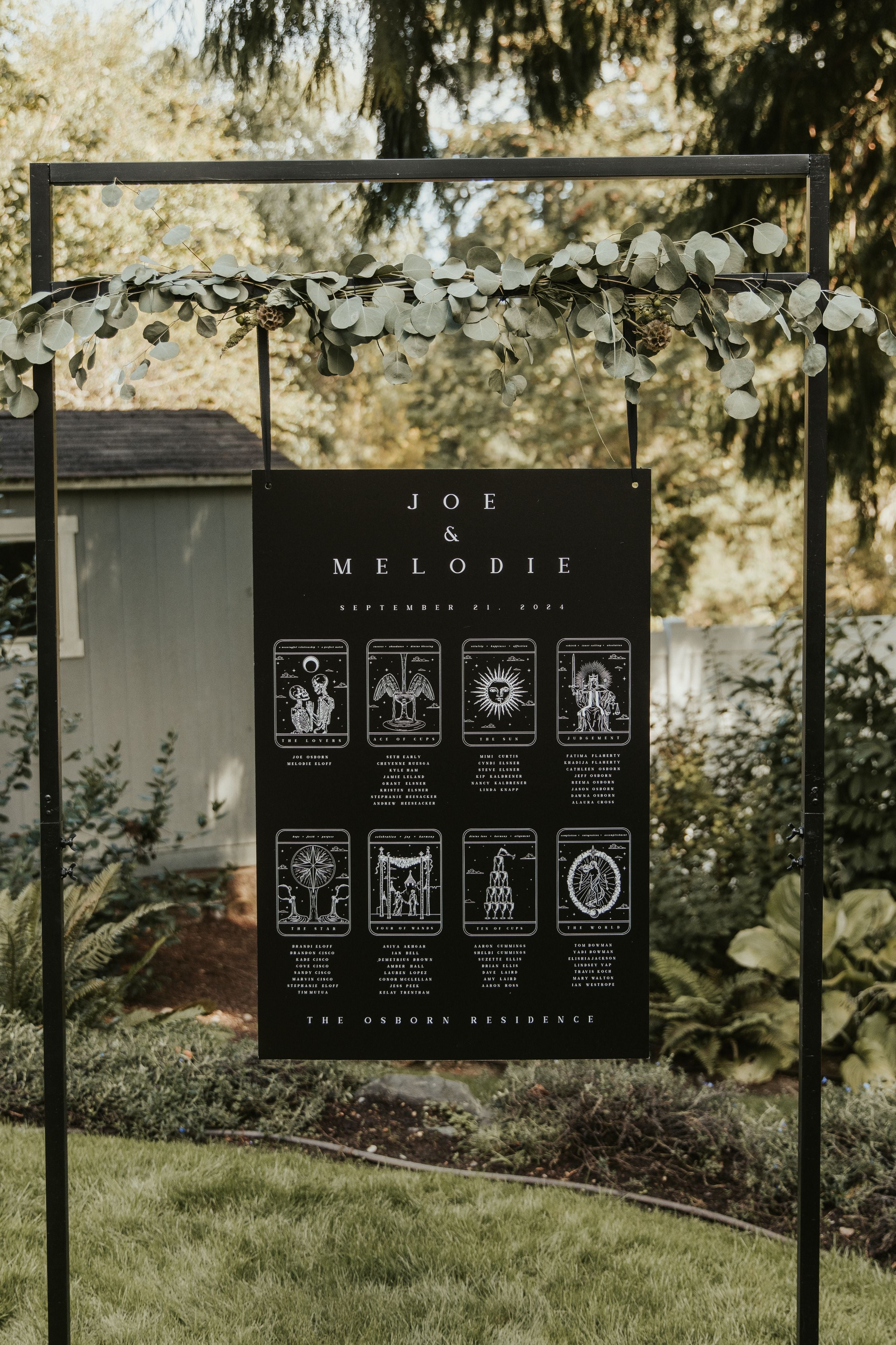 A luxury black wedding sign with gothic white illustrations hangs from a metal stand with greenery in an elegant UK garden setting.