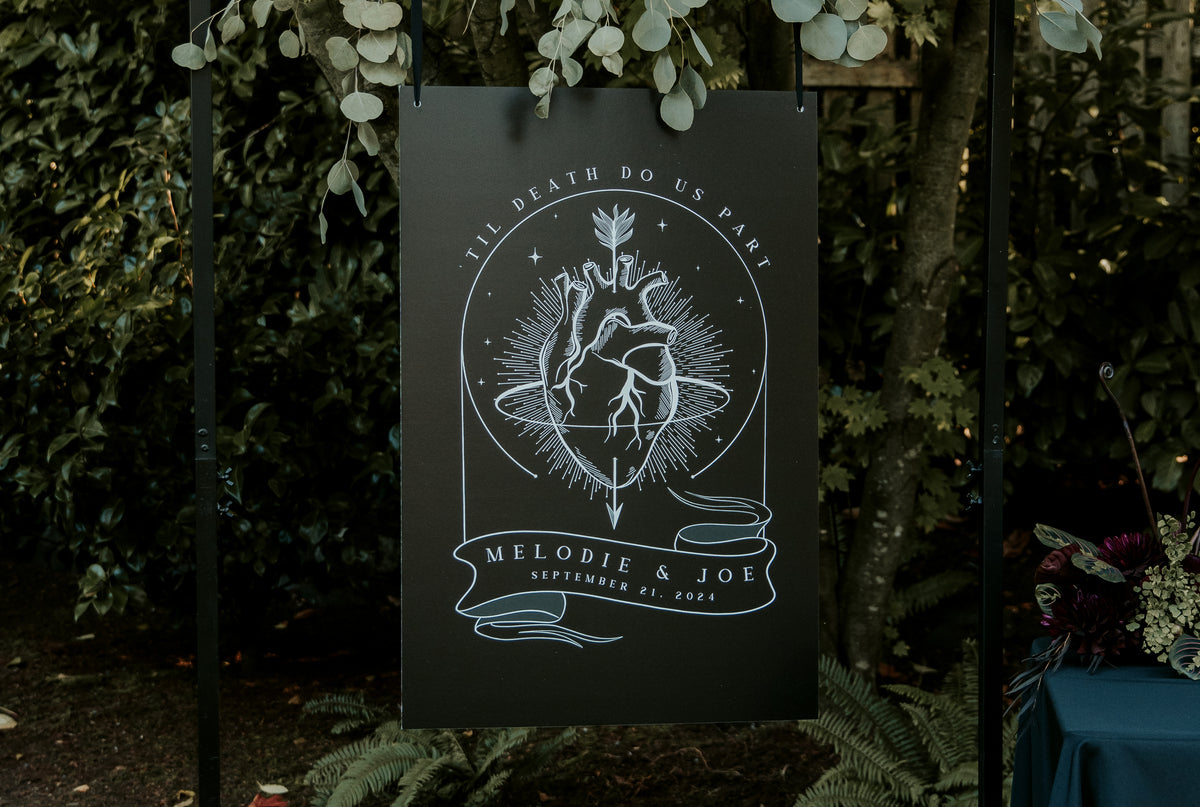 A gothic wedding welcome sign by Birdhouse197 with deep red and black design hangs outdoors among lush greenery.