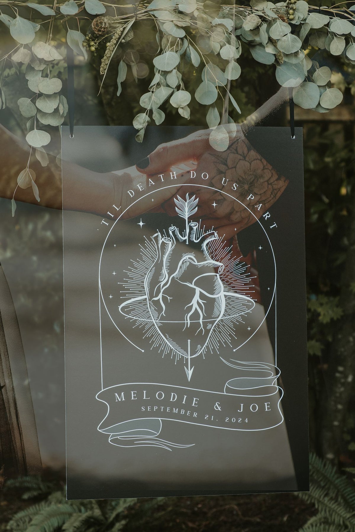 A dark romantic wedding welcome sign with botanical illustrations and anatomical hearts, set against a glass background reflecting greenery.