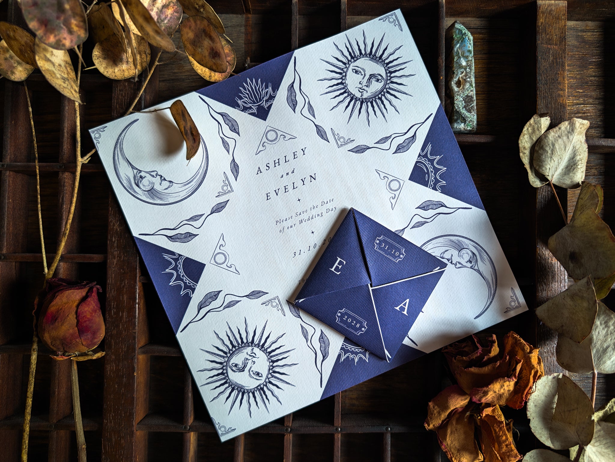 A luxury celestial-themed wedding stationery sample pack is elegantly arranged on a wooden surface, featuring invitations with delicate sun, moon, and star motifs. The design exudes a mystical and romantic feel, complemented by dried flowers, foliage, and a vintage-inspired decorative stone for an alternative UK boho aesthetic.