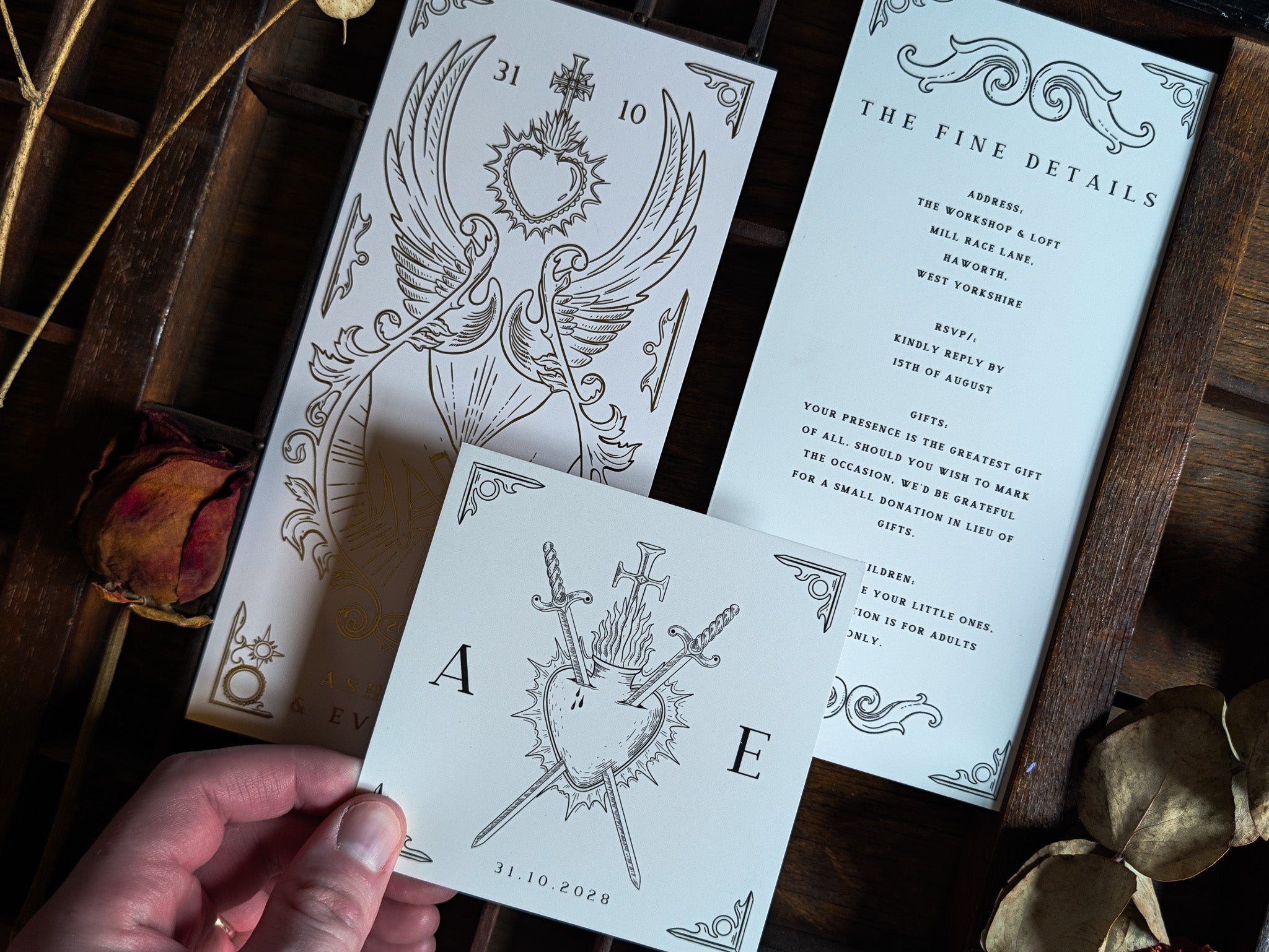 A hand displays a premium wedding invitation card with a striking tarot-inspired illustration of swords and a heart, above two ornate, gothic wedding stationery pieces featuring dramatic winged heart motifs. The luxurious suite is styled with dried roses and leaves arranged on a vintage dark wooden surface, creating an alternative and romantic UK setting.