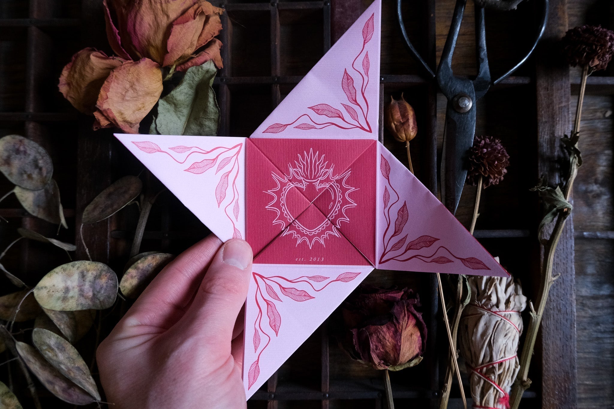 A hand presents luxury origami-style celestial wedding stationery in pink and red, amid dried flowers and herbs on rustic wood.