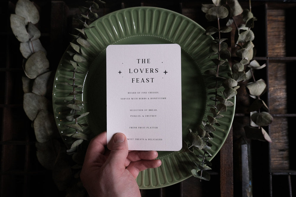 Hand holding a Bleeding Heart Lovers Feast menu with tarot style art over a green plate