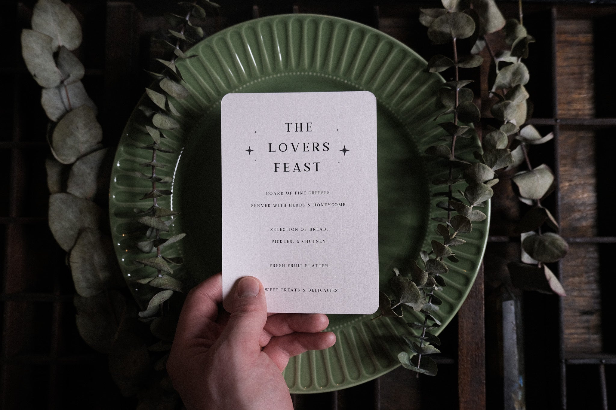 A hand holds luxury gothic wedding menu stationery with mystical tarot-inspired artwork, displayed over a green plate with dried leaves.