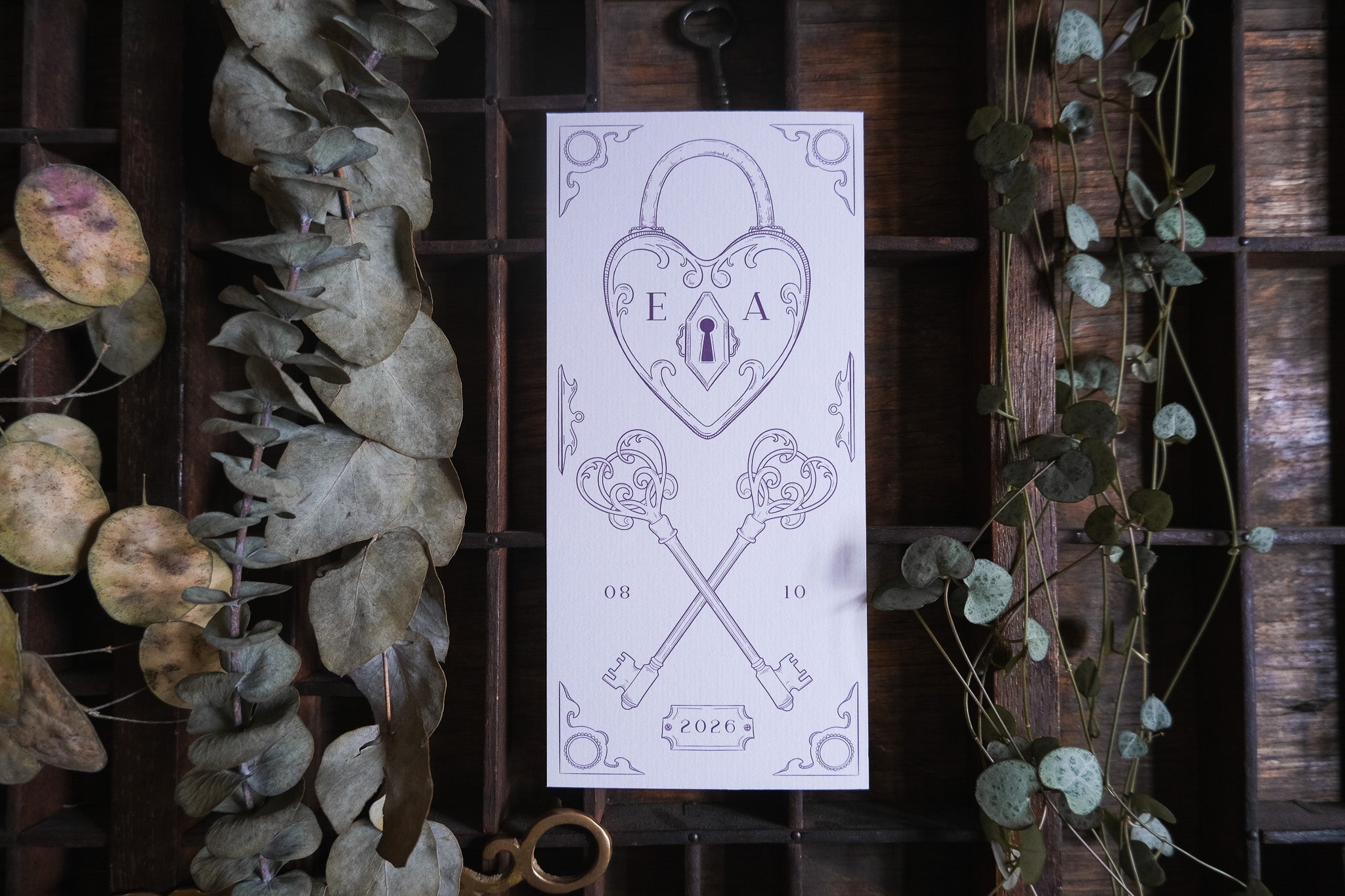 Luxury vintage save the date card with a heart padlock, crossed keys and dried foliage on rustic wood—romantic UK wedding stationery.