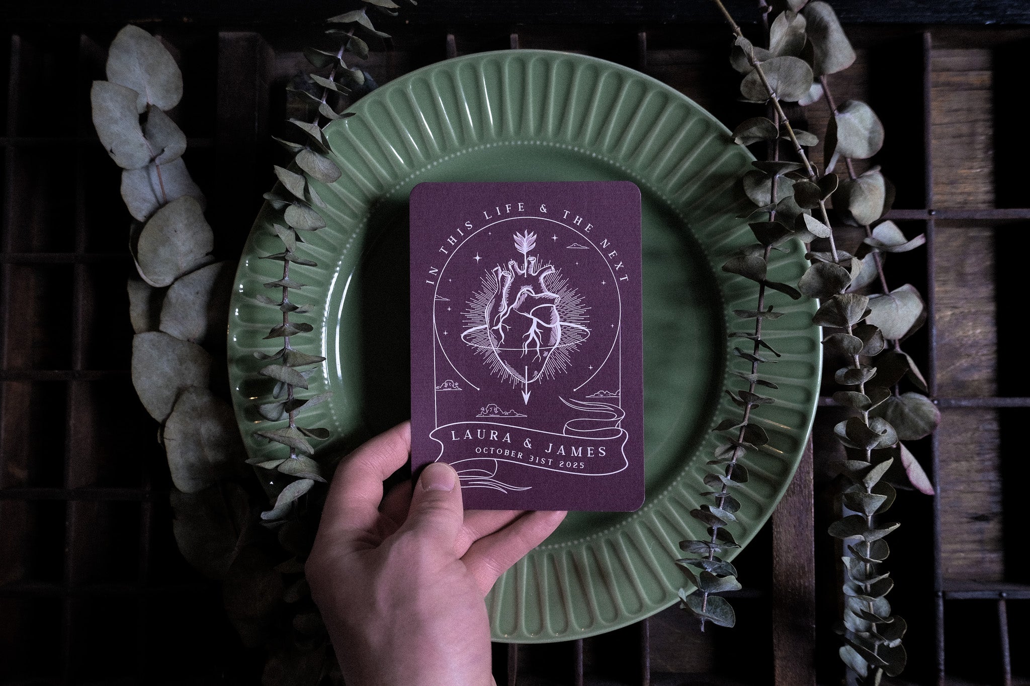 A hand displays a gothic maroon wedding menu card with vintage anatomical heart art, luxury cardstock, on a green plate amid dried leaves.