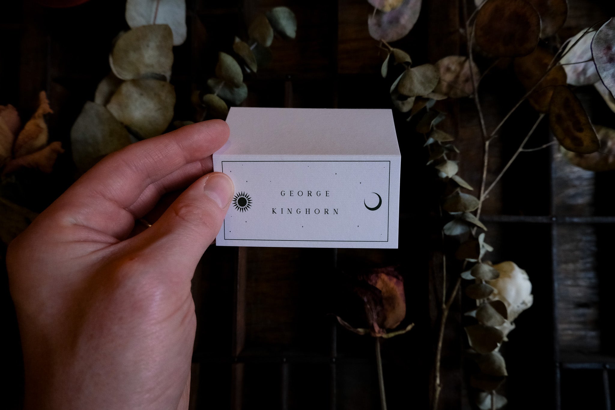 A hand displays a celestial-themed luxury wedding placecard with sun and moon icons, set against dried florals for a romantic gothic look.